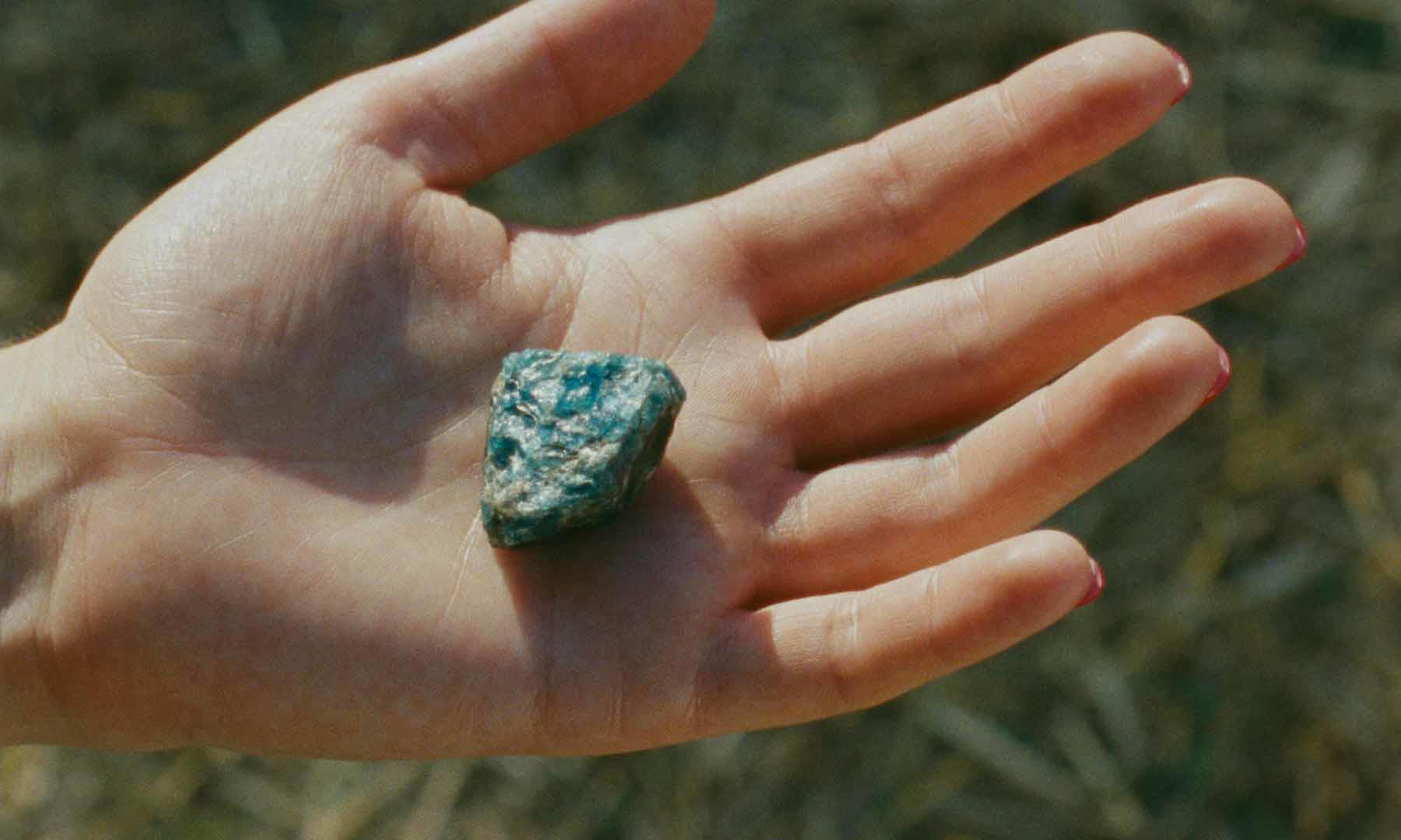 Eine Hand hält einen blaugrünen Stein. Die Hand hat rosa Nagellack. Der Hintergrund ist ein verschwommenes Feld.