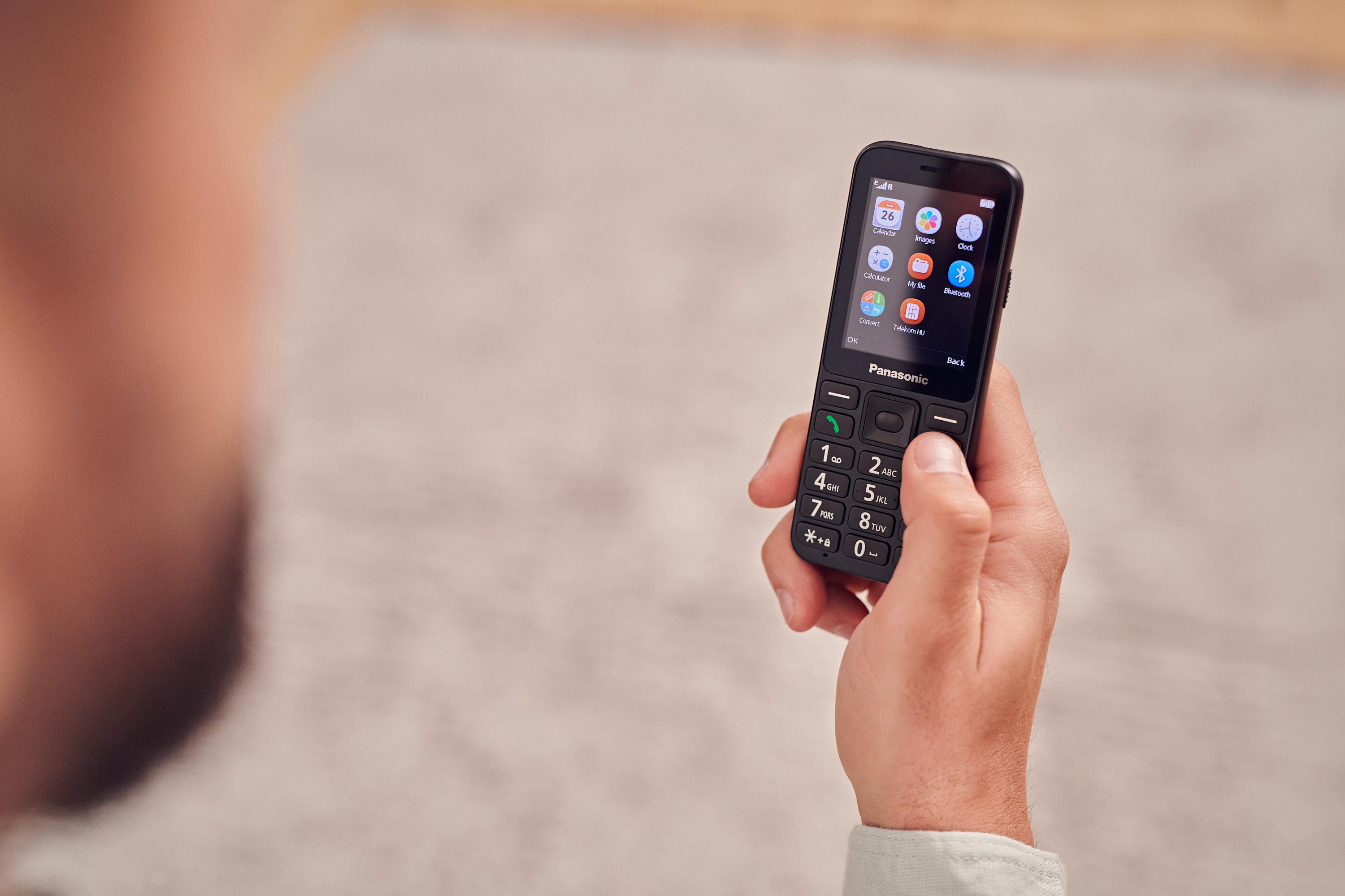 Eine Hand hält ein schwarzes Panasonic-Telefon. Der Bildschirm zeigt Symbole für Kalender, Bilder, Uhr, Taschenrechner und Bluetooth.