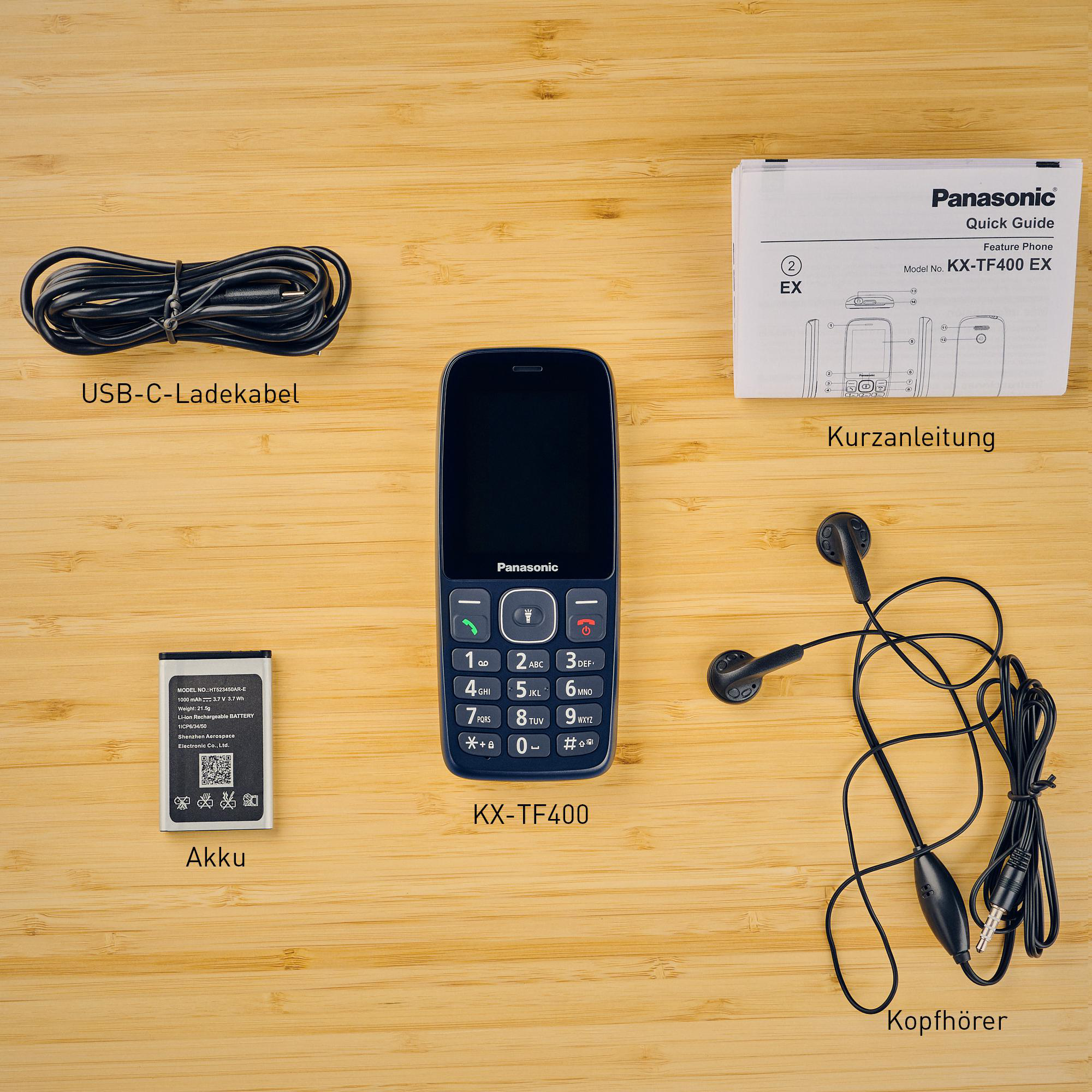 Ein Panasonic KX-TF400-Telefon mit Zubehör wie Ladegerät, Kopfhörern, Akku und Handbuch auf Holzoberfläche.