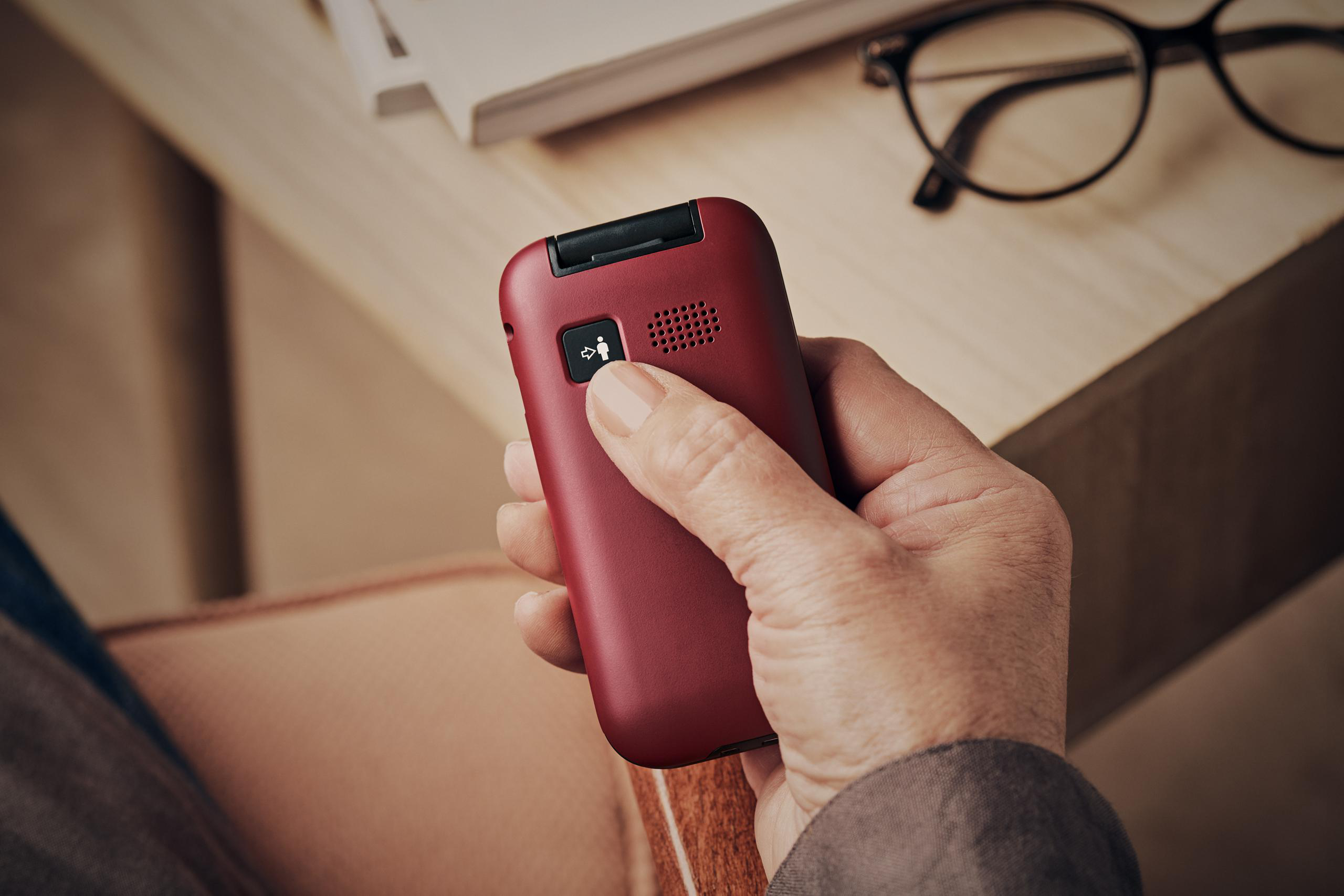 Eine Hand hält ein rotes Klapphandy. Ein Knopf mit einer Person und einem Pfeil ist sichtbar. Brille und Buch im Hintergrund.