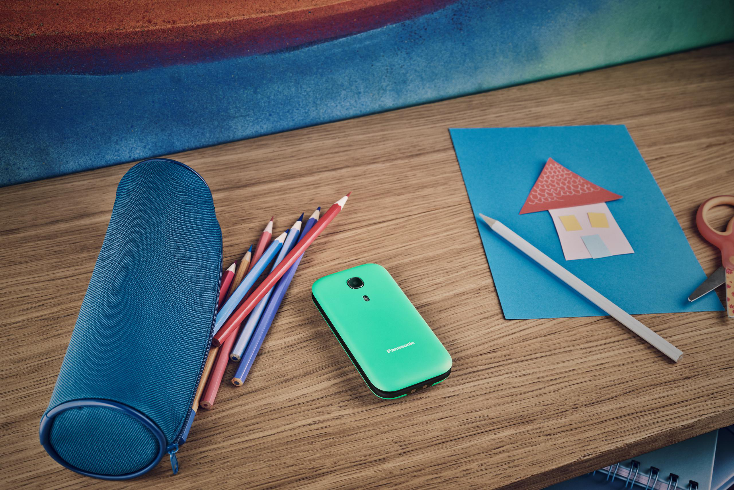Holztisch mit blauem Federmäppchen, Bleistiften, grünem Telefon und Papierschnitthaus.
