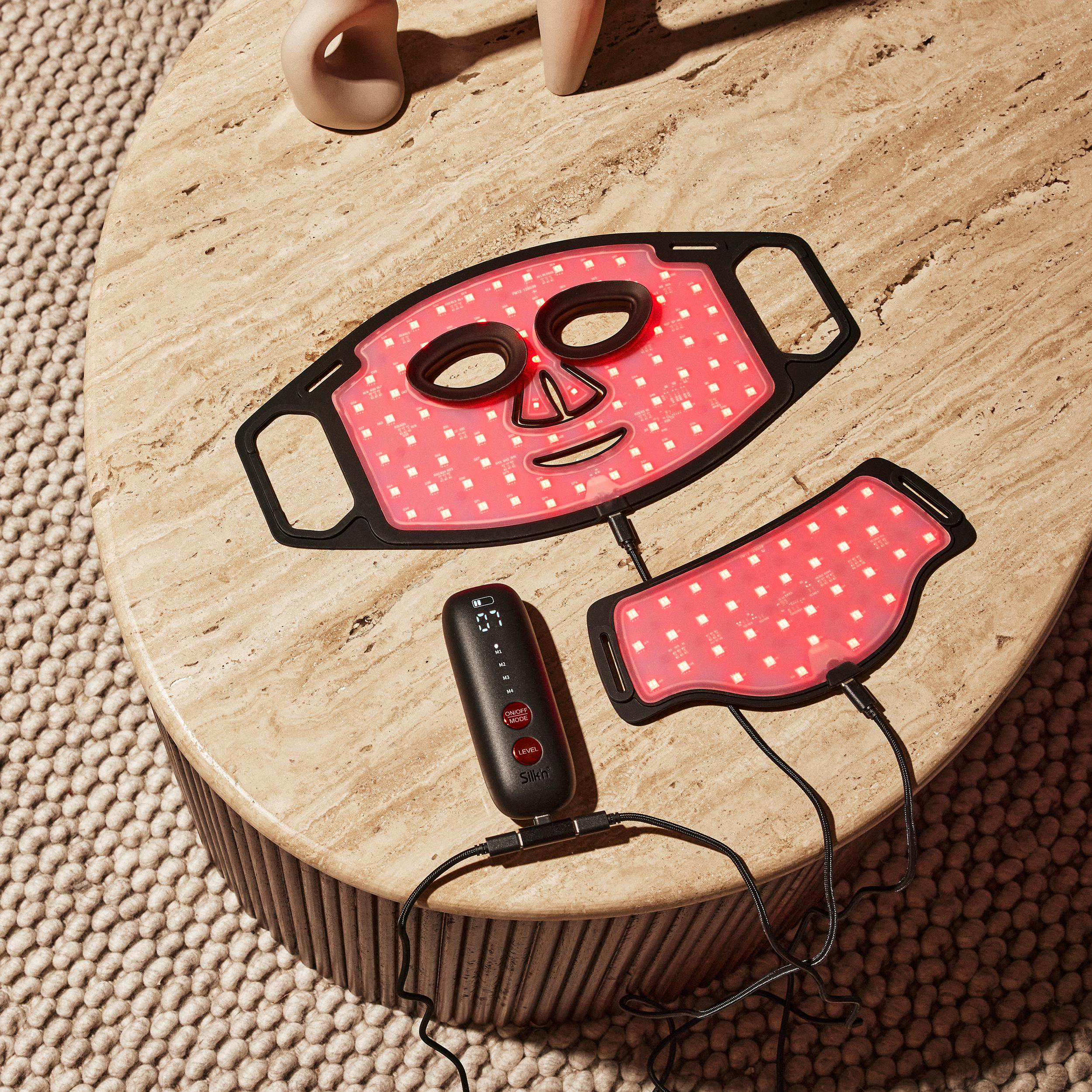 LED-Gesichtsmaske und Nackenteil auf einem Marmortisch mit Fernbedienung. Beiger Teppich im Hintergrund.