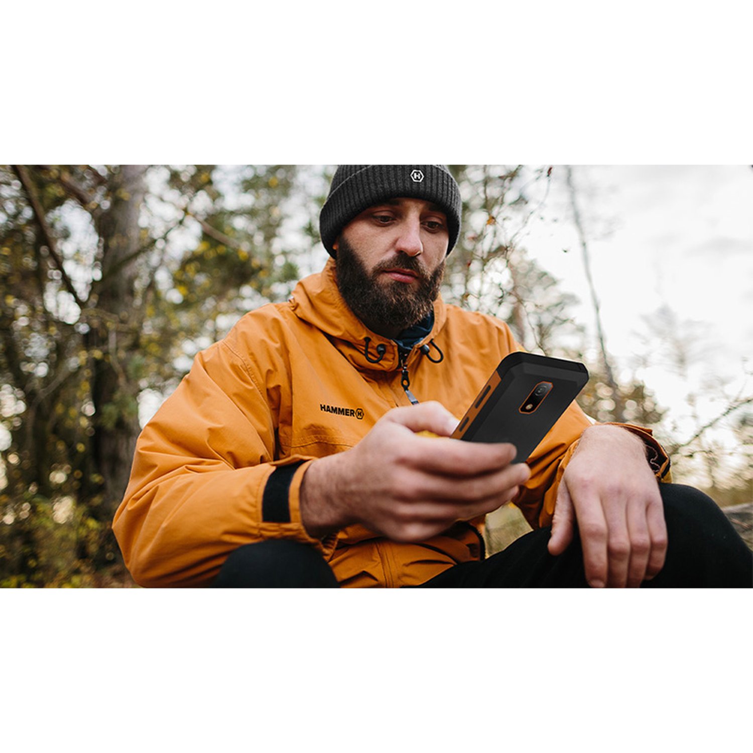 Man met muts en oranje jas kijkt naar telefoon. Bosachtergrond, bewolkte lucht. Hammer-logo.