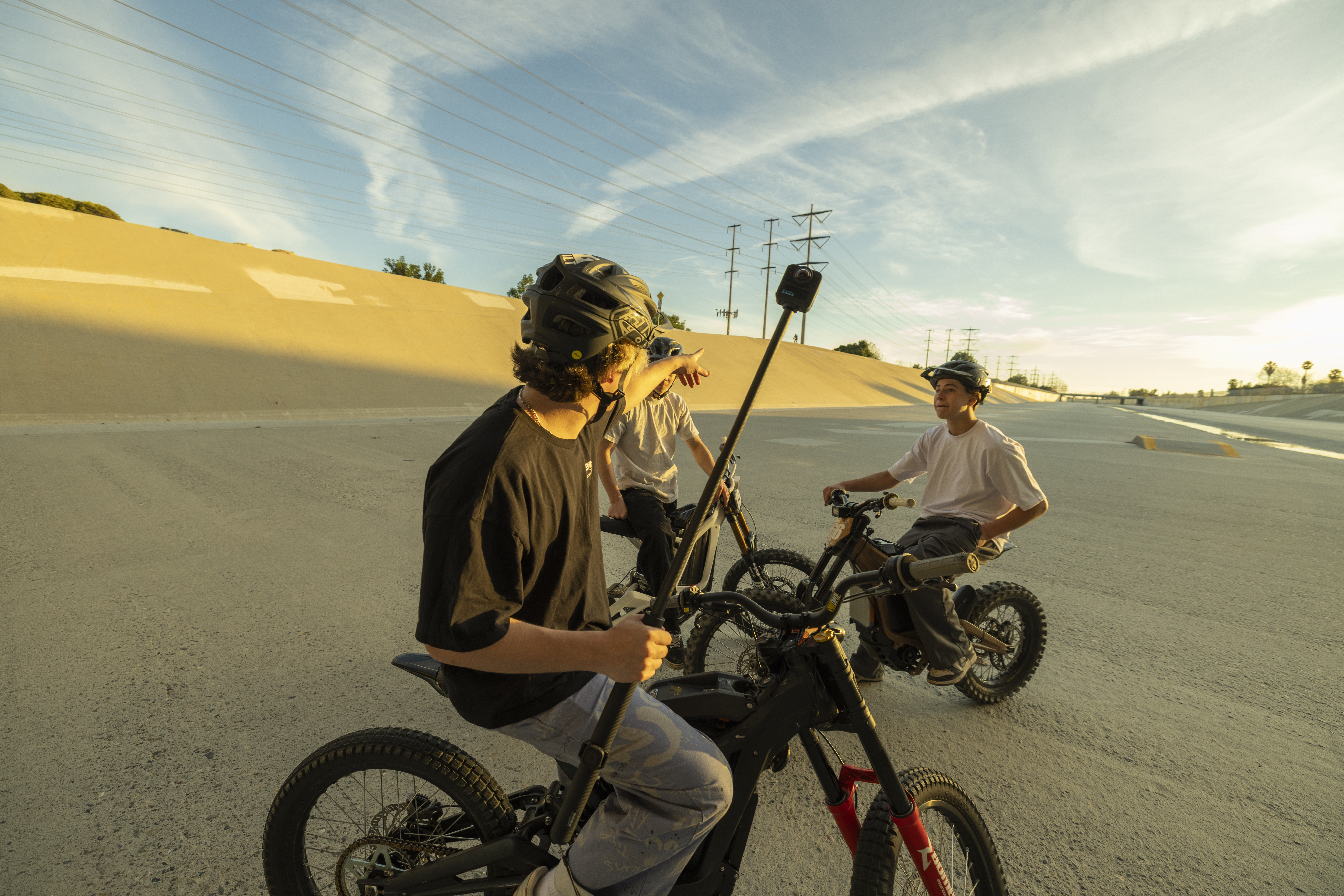Drei Personen auf E-Bikes. Einer zeigt in den Himmel. Hinter ihnen befindet sich eine Betonkonstruktion. Der Himmel ist blau.