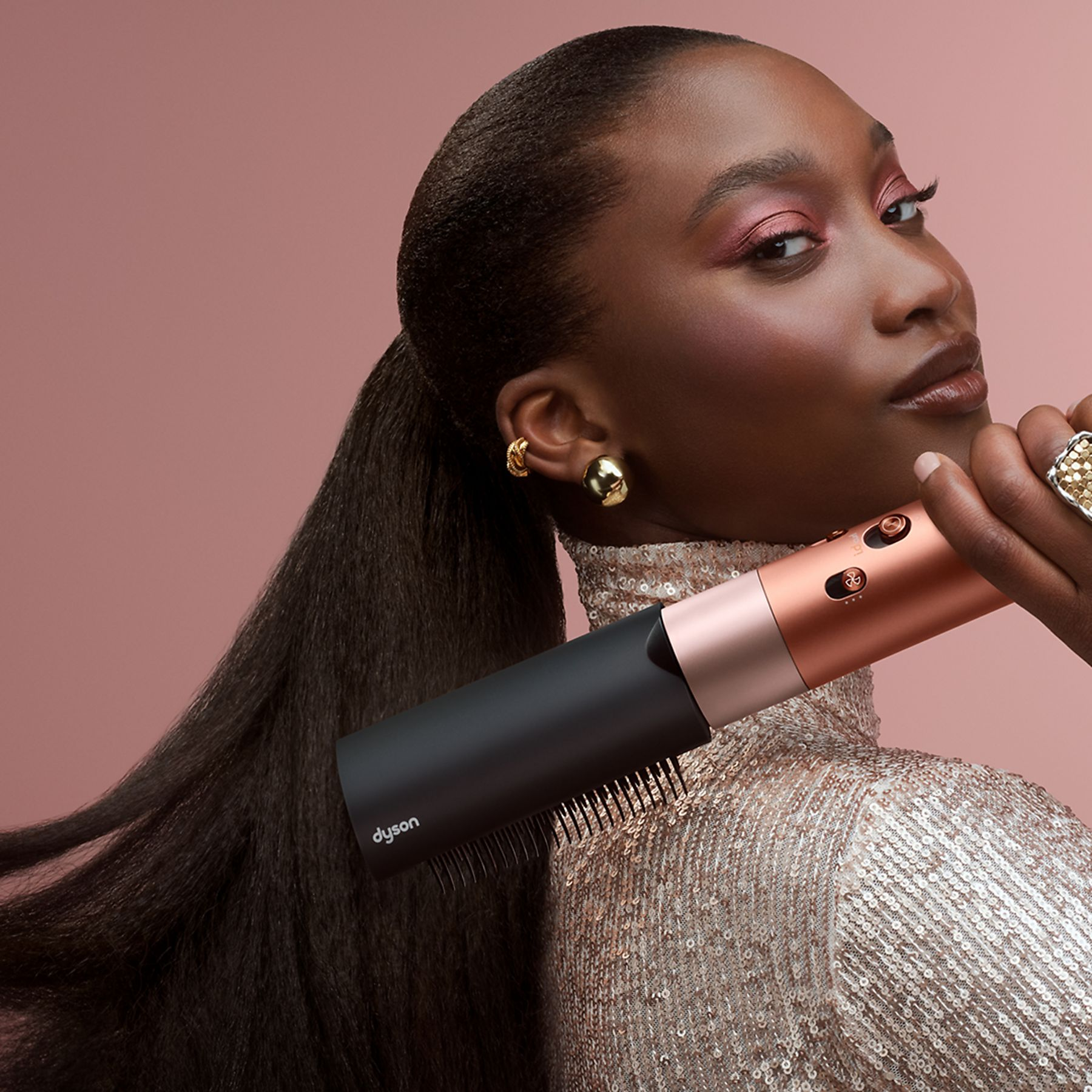 Kobieta trzyma narzędzie do stylizacji włosów Dyson. Ma ciemną skórę, nosi złote kolczyki i top z cekinami. Różowe tło.