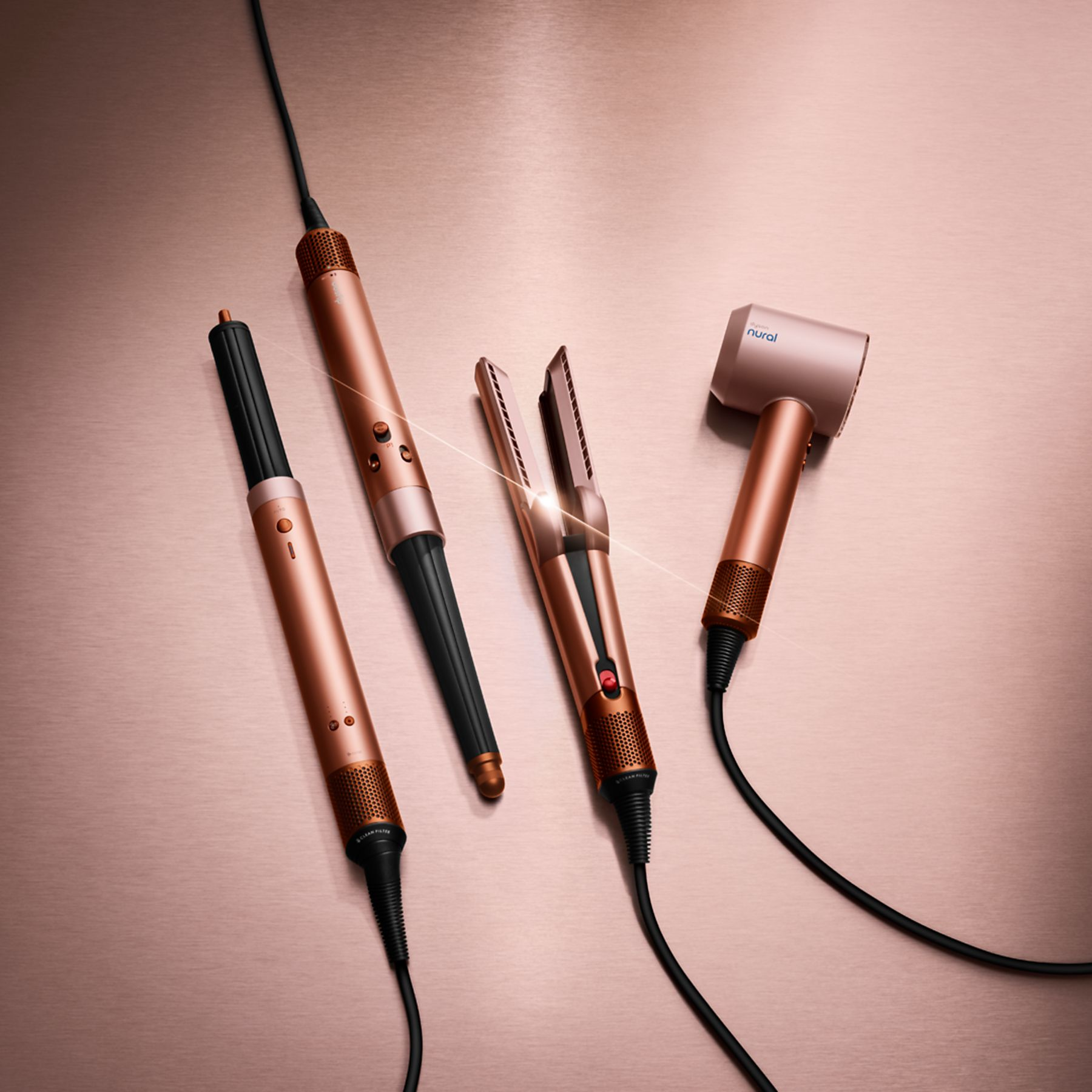 Cztery narzędzia do stylizacji włosów w kolorze różowego złota na różowej metalicznej powierzchni. Zawiera suszarkę, prostownicę i lokówki z czarnymi kablami.