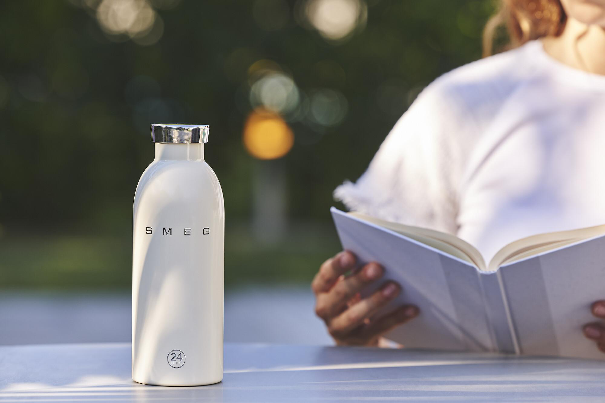Eine weiße Wasserflasche steht auf einem Tisch. Im Hintergrund liest eine Person ein Buch. Grüner Bokeh-Hintergrund.