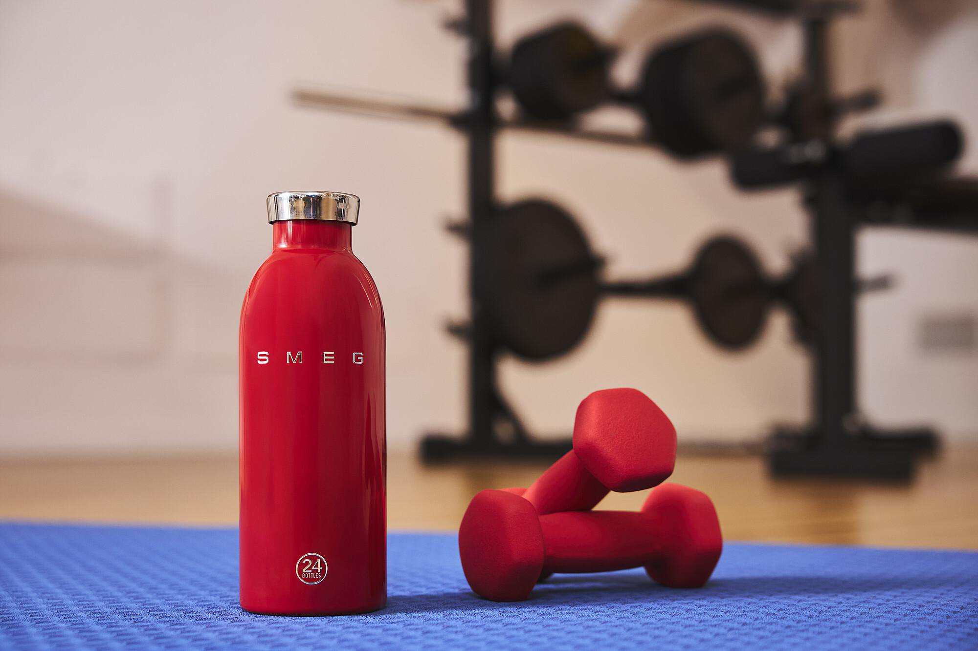Rote Wasserflasche und Hanteln auf blauer Matte. Gewichtsregal im verschwommenen Hintergrund. Gym-Szene.