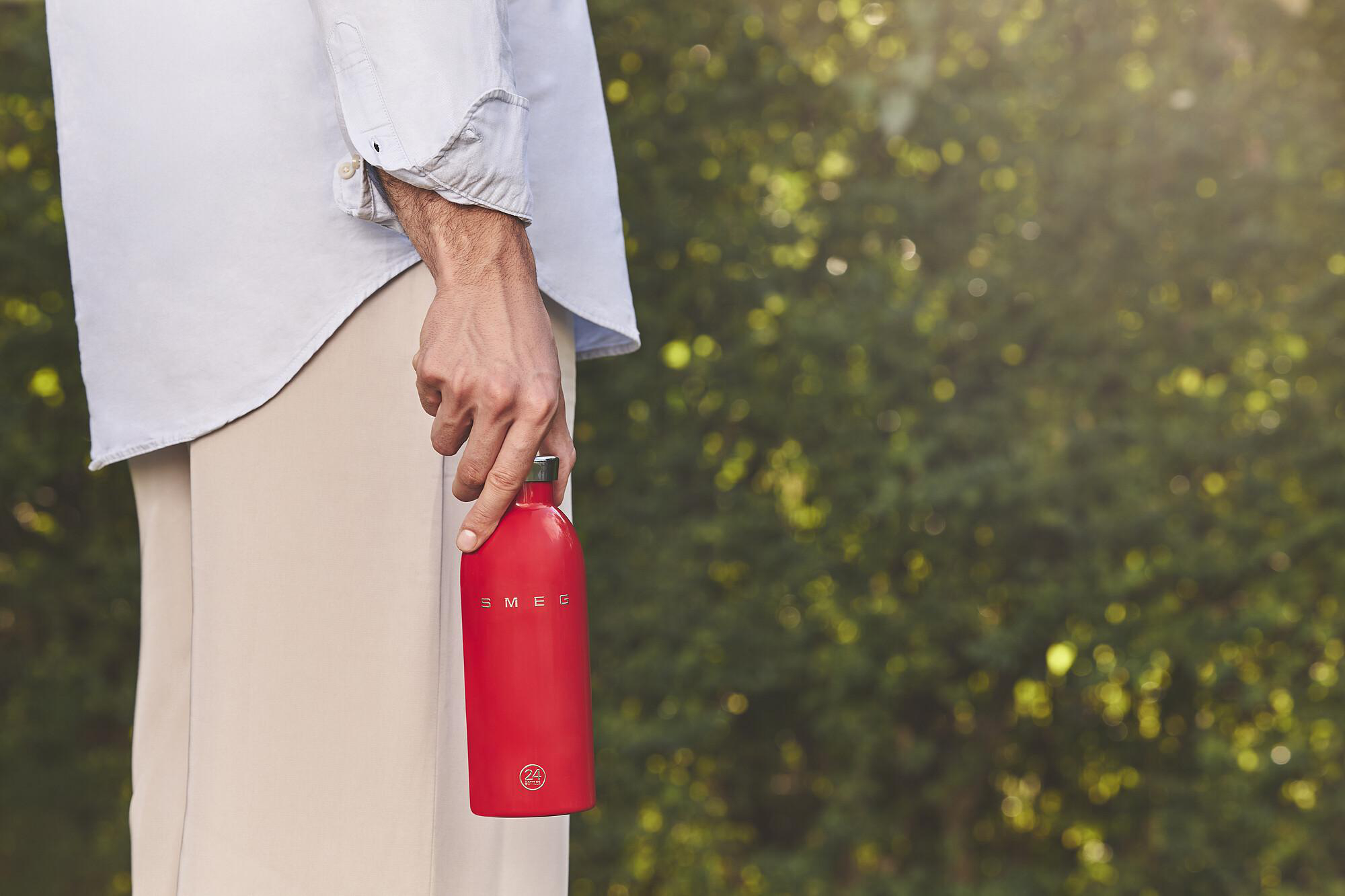 Eine Person hält eine rote Wasserflasche. Sie trägt ein hellblaues Hemd und beige Hosen. Grünes Laub ist im Hintergrund.