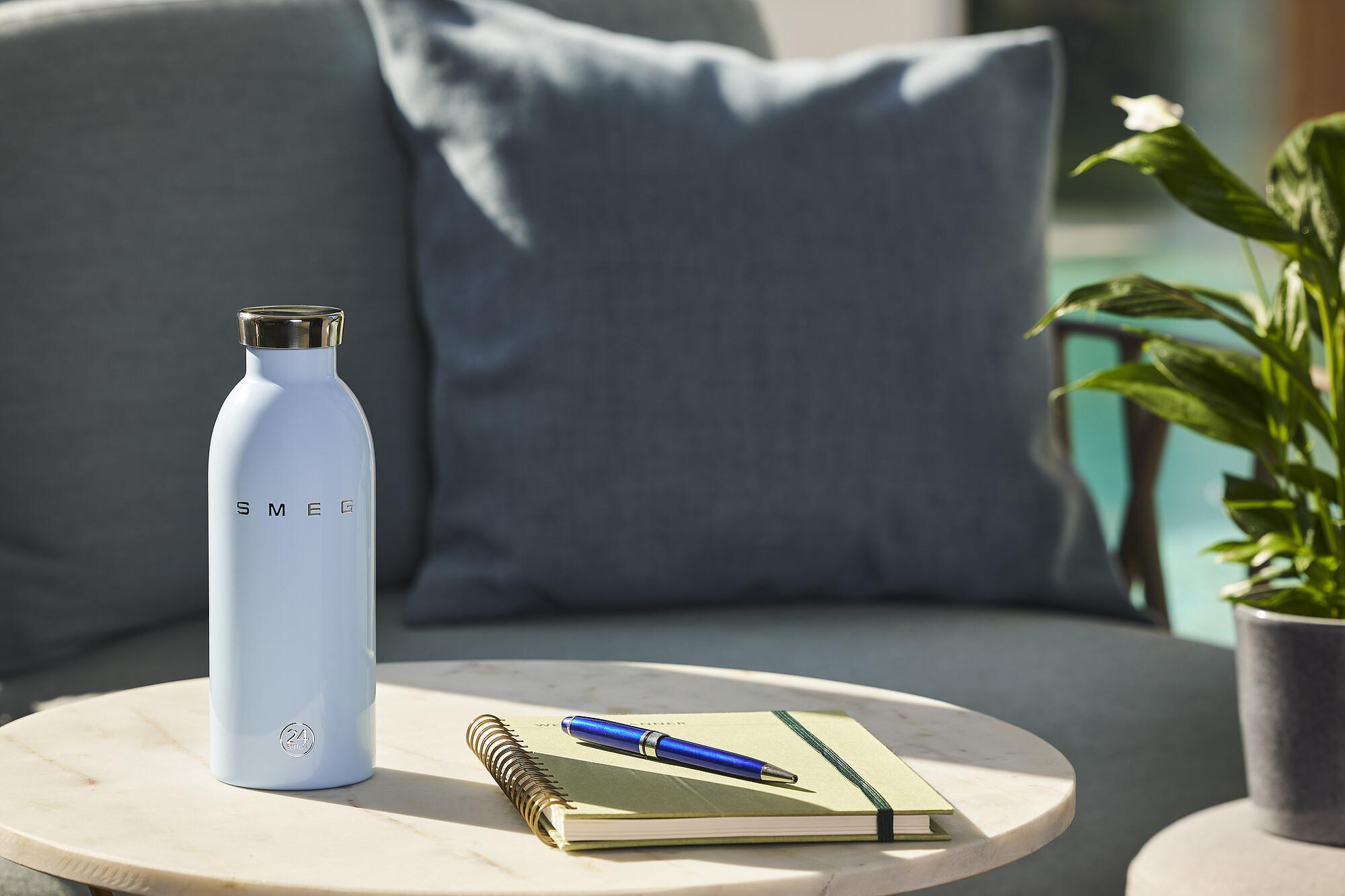 Eine hellblaue SMEG-Wasserflasche, Stift, Notizbuch auf einem Tisch. Eine Couch mit Kissen und Topfpflanze im Hintergrund.
