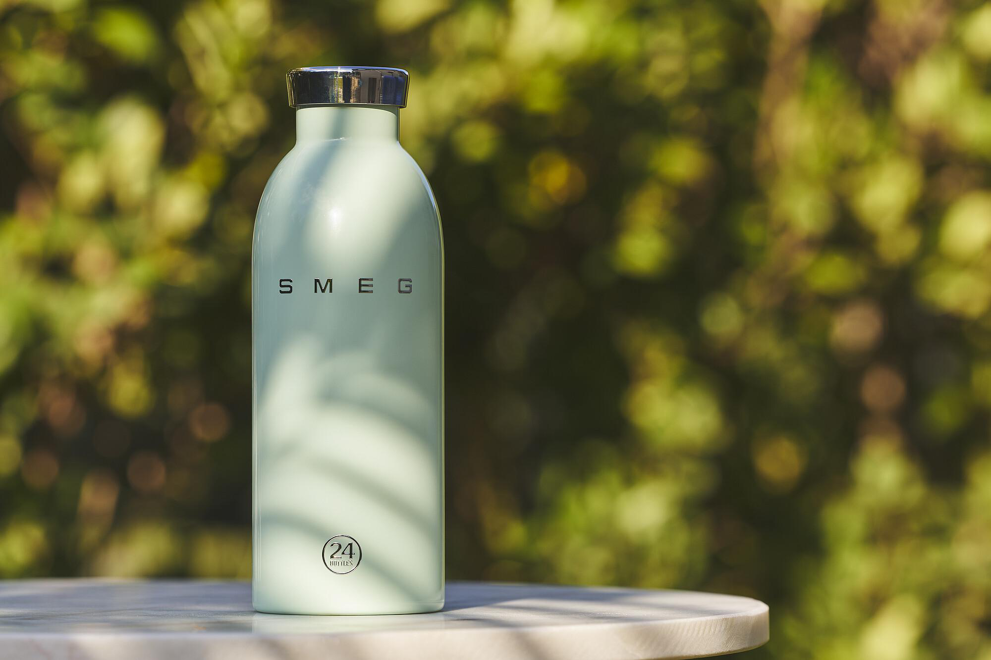 Eine hellgrüne SMEG-Wasserflasche steht auf einem Tisch. Der Hintergrund ist ein verschwommenes grünes Laub.