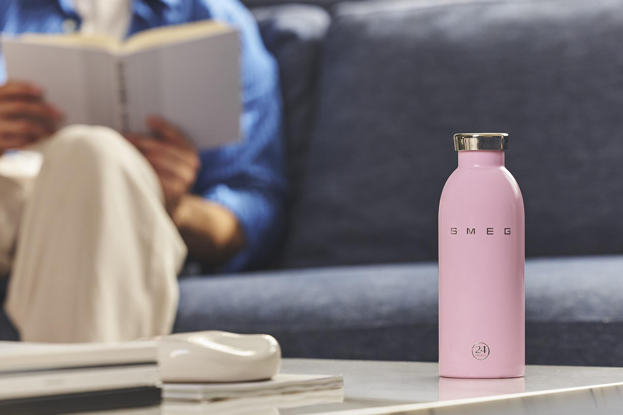 Rosa SMEG-Wasserflasche auf einem Tisch. Im Hintergrund liest eine Person ein Buch auf einer Couch.