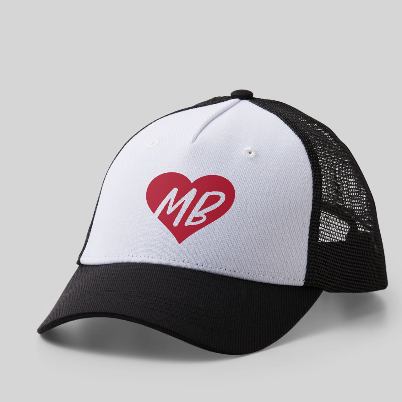 Eine weiß-schwarze Trucker-Mütze. Auf der Vorderseite befindet sich ein rotes Herz mit weißen Buchstaben "MB". Der Hintergrund ist grau.