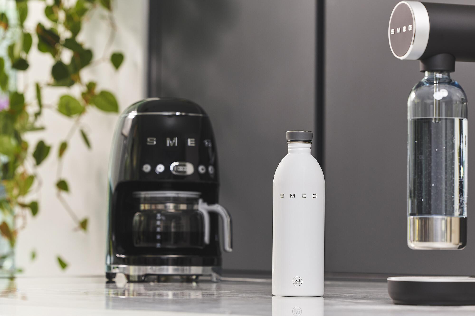 Eine Küchenszene mit einer schwarzen Kaffeemaschine, einer weißen Wasserflasche und einem Wasserspender. Grüne Blätter im Hintergrund.