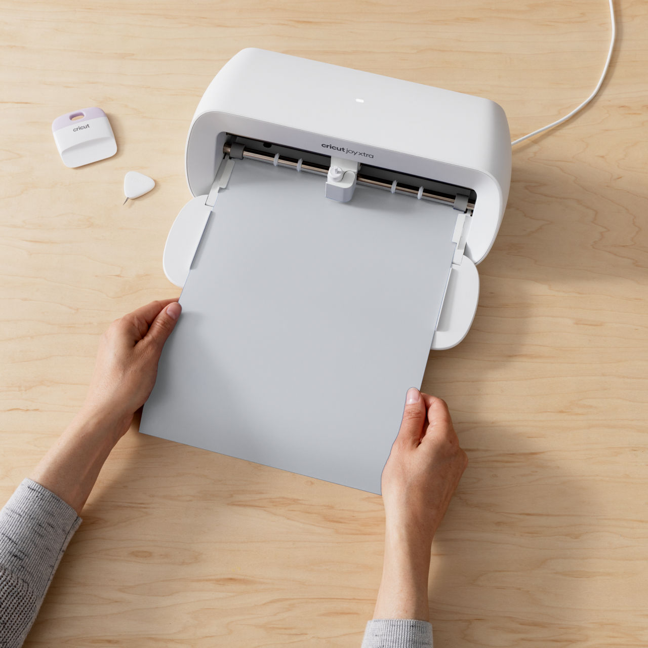 Hände halten ein graues Blatt vor einer weißen Cricut-Maschine auf einer Holzoberfläche. Cricut-Werkzeuge sind links.