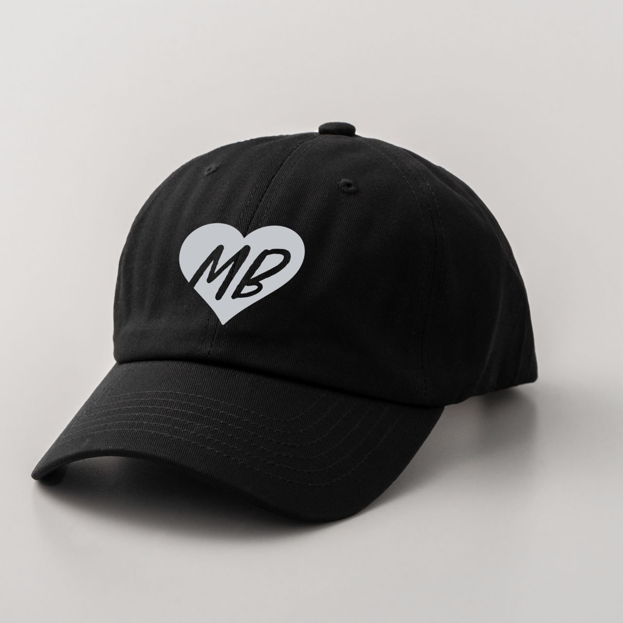 Eine schwarze Baseballkappe mit einem weißen Herz mit 'MB' darin. Die Kappe befindet sich auf weißem Hintergrund.