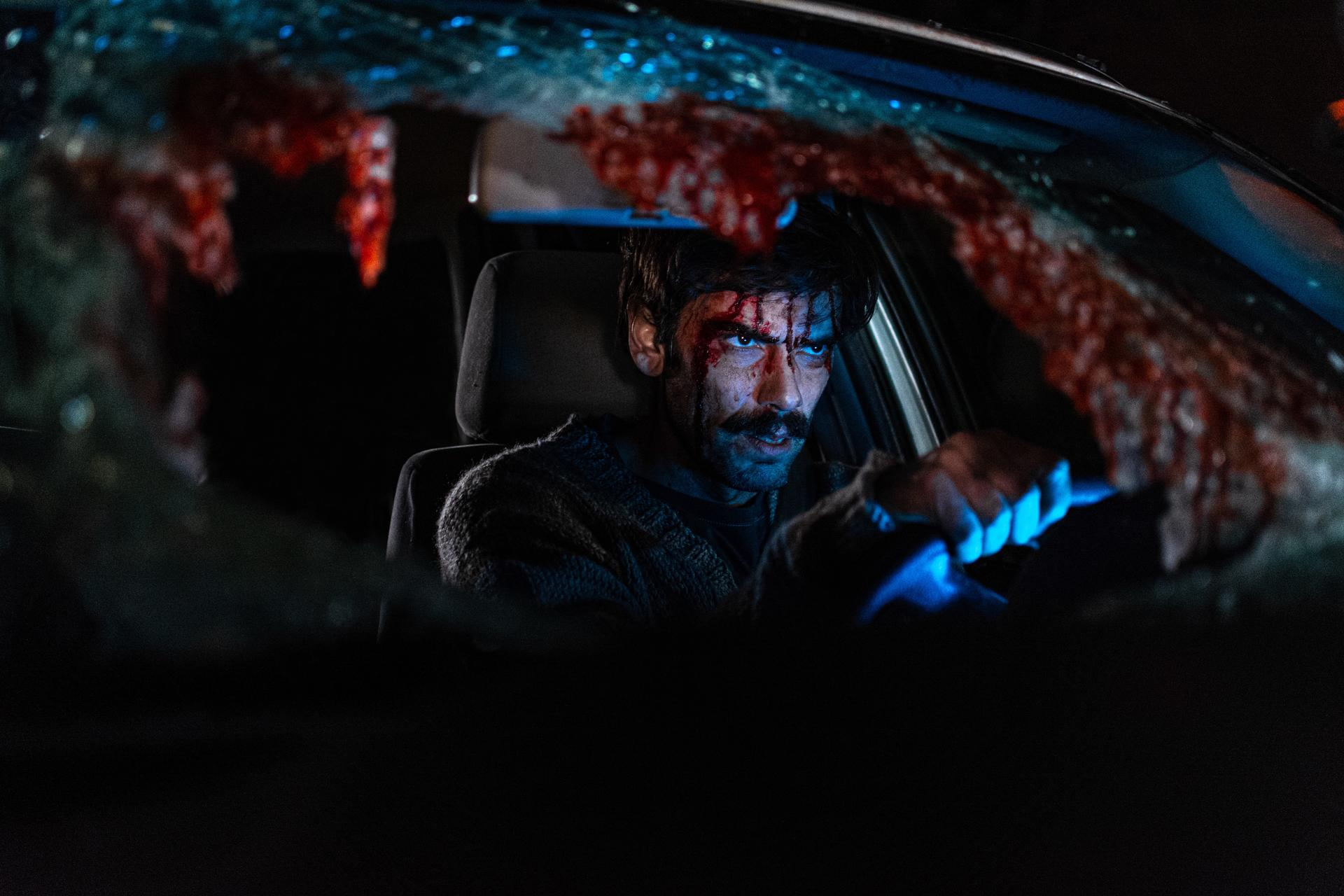 Mann mit Blut im Gesicht im Auto. Zersplitterte Windschutzscheibe mit Blut. Dunkle Szene, intensiver Ausdruck.