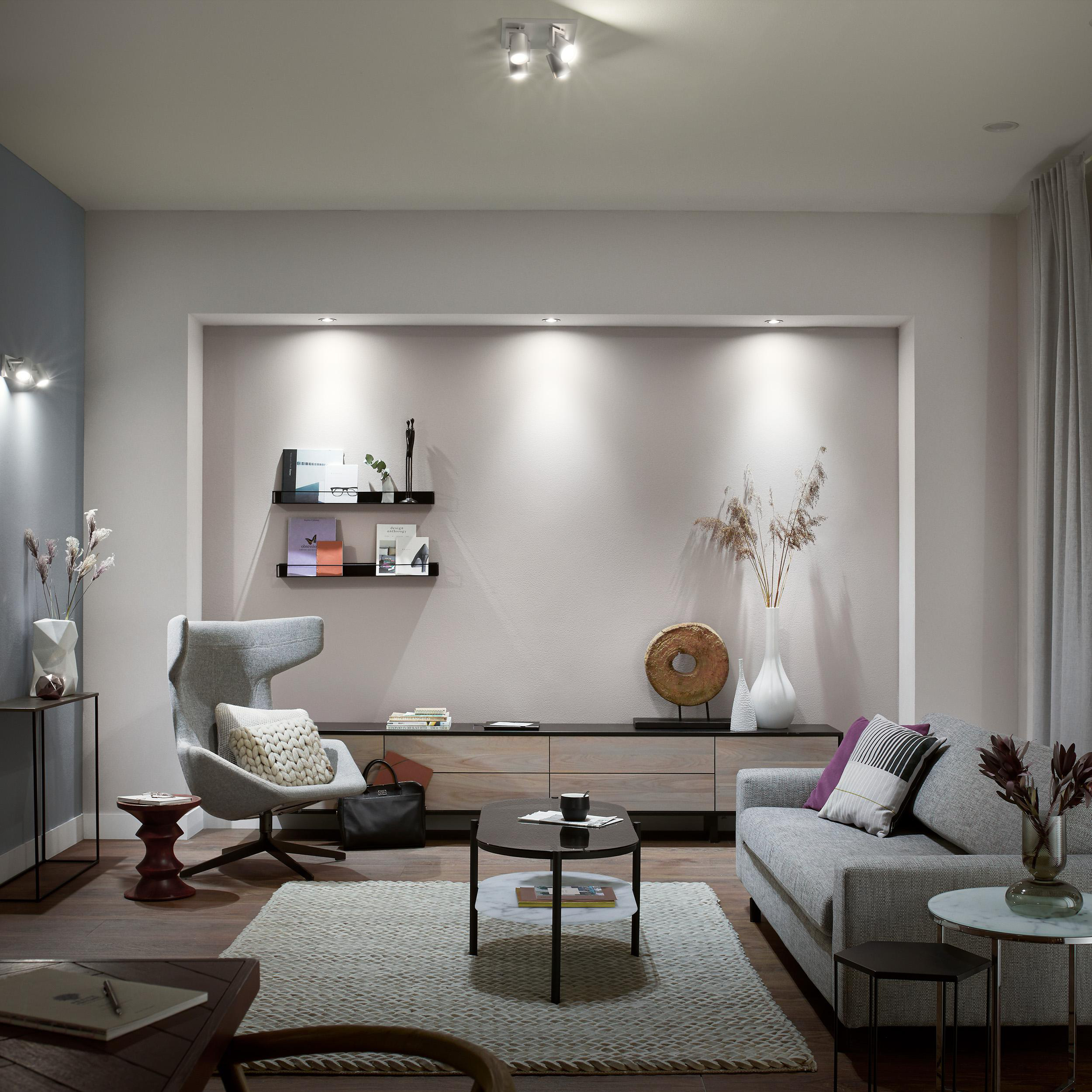 Wohnzimmer mit Sofa, Sessel, Couchtisch, Teppich und Dekor. Neutrale Farben dominieren den Raum. Lichter sind an.