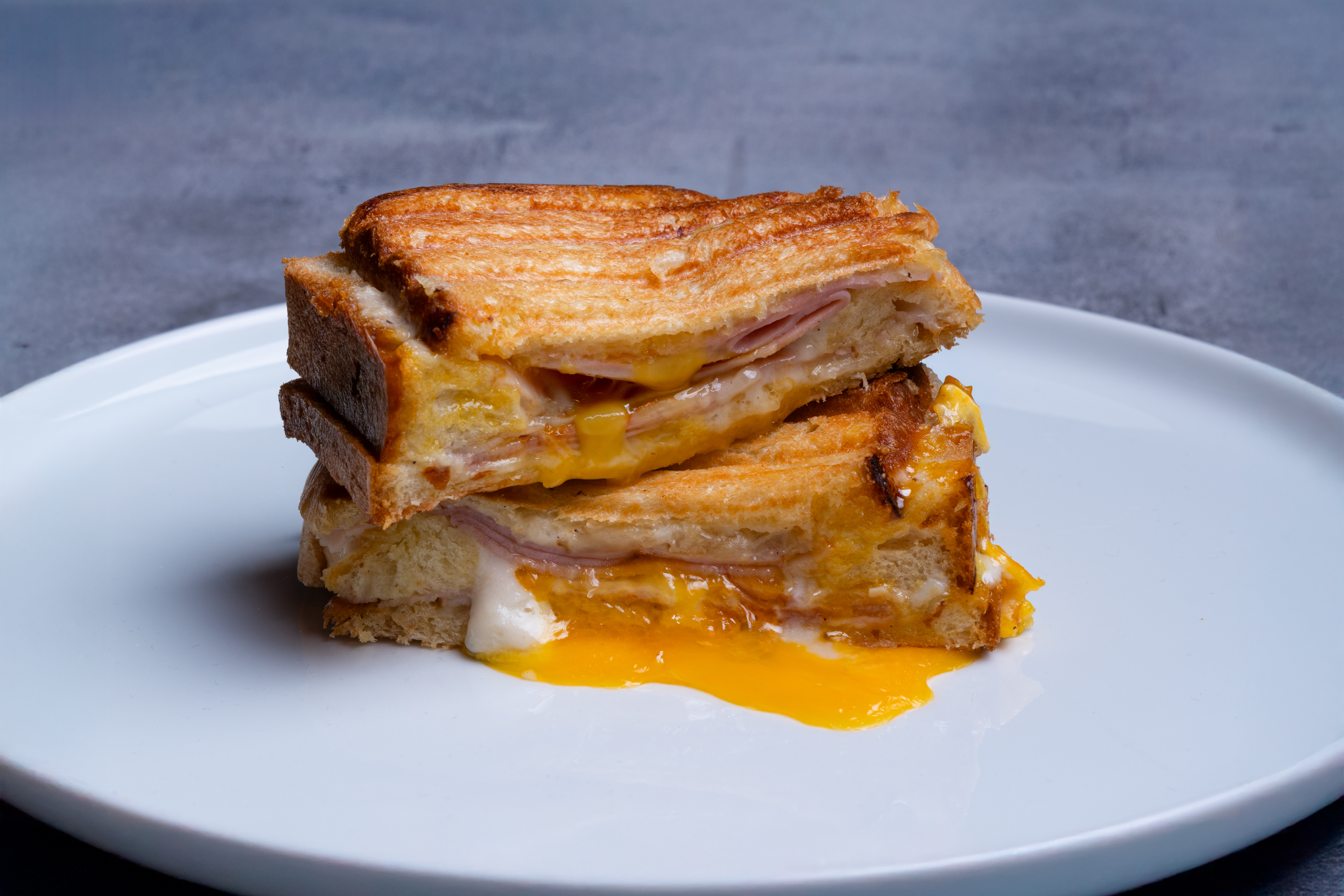 Ein geschichteter gegrillter Käsesandwich mit Schinken auf einem weißen Teller. Geschmolzener Käse und Eigelb sind sichtbar.