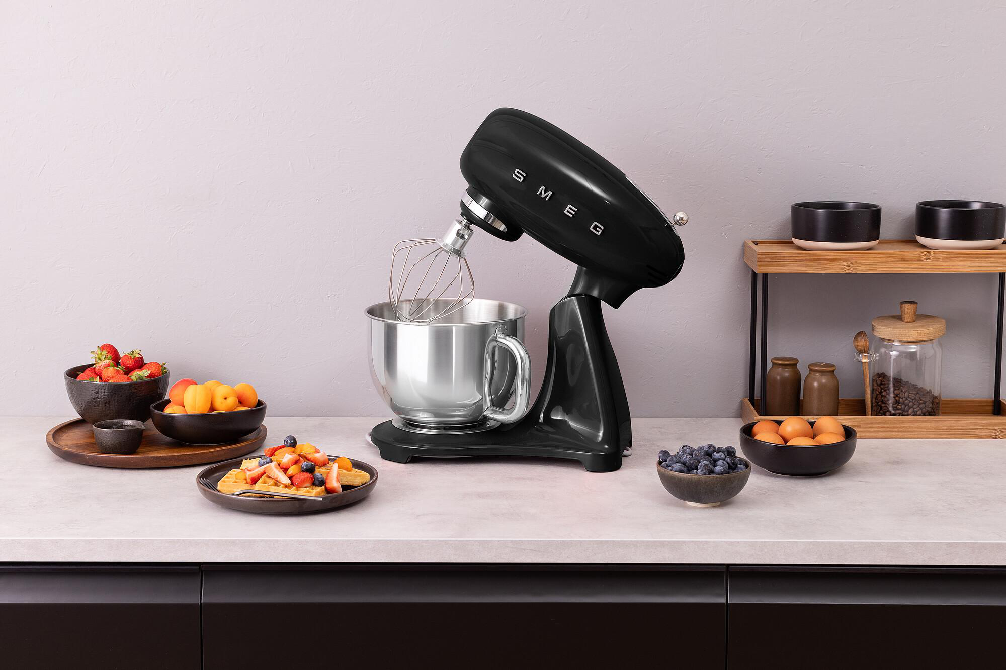 Schwarze SMEG-Küchenmaschine mit Schneebesen, Früchten und Waffeln. Schalen mit Beeren und Eiern. Holzregal mit Gläsern und Schalen.