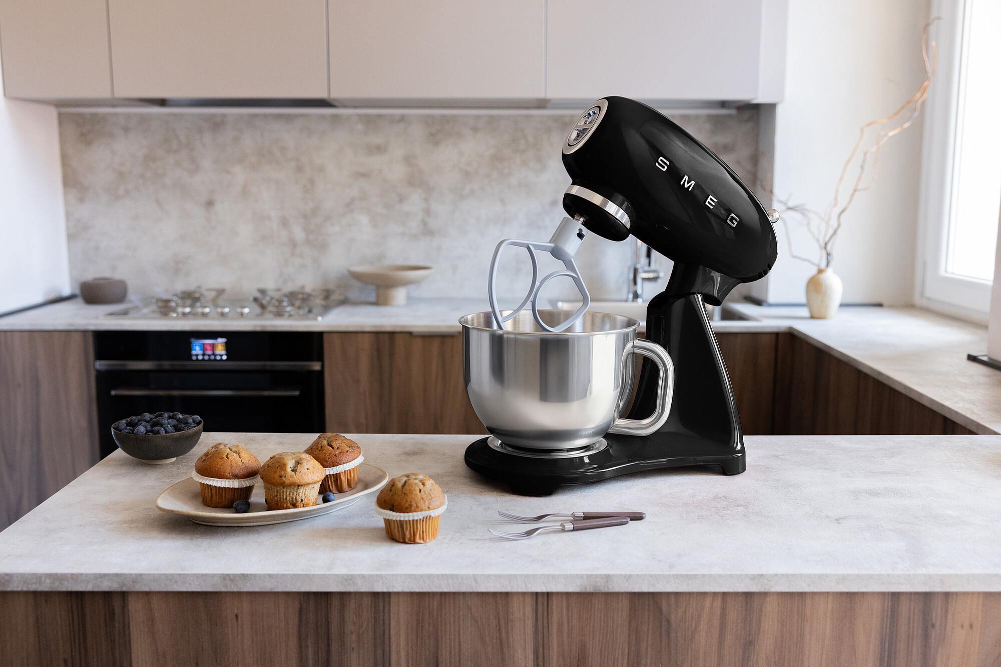 Ein schwarzer Küchenmixer mit silberner Schüssel und Schneebesen. Muffins und Blaubeeren liegen auf der Theke.