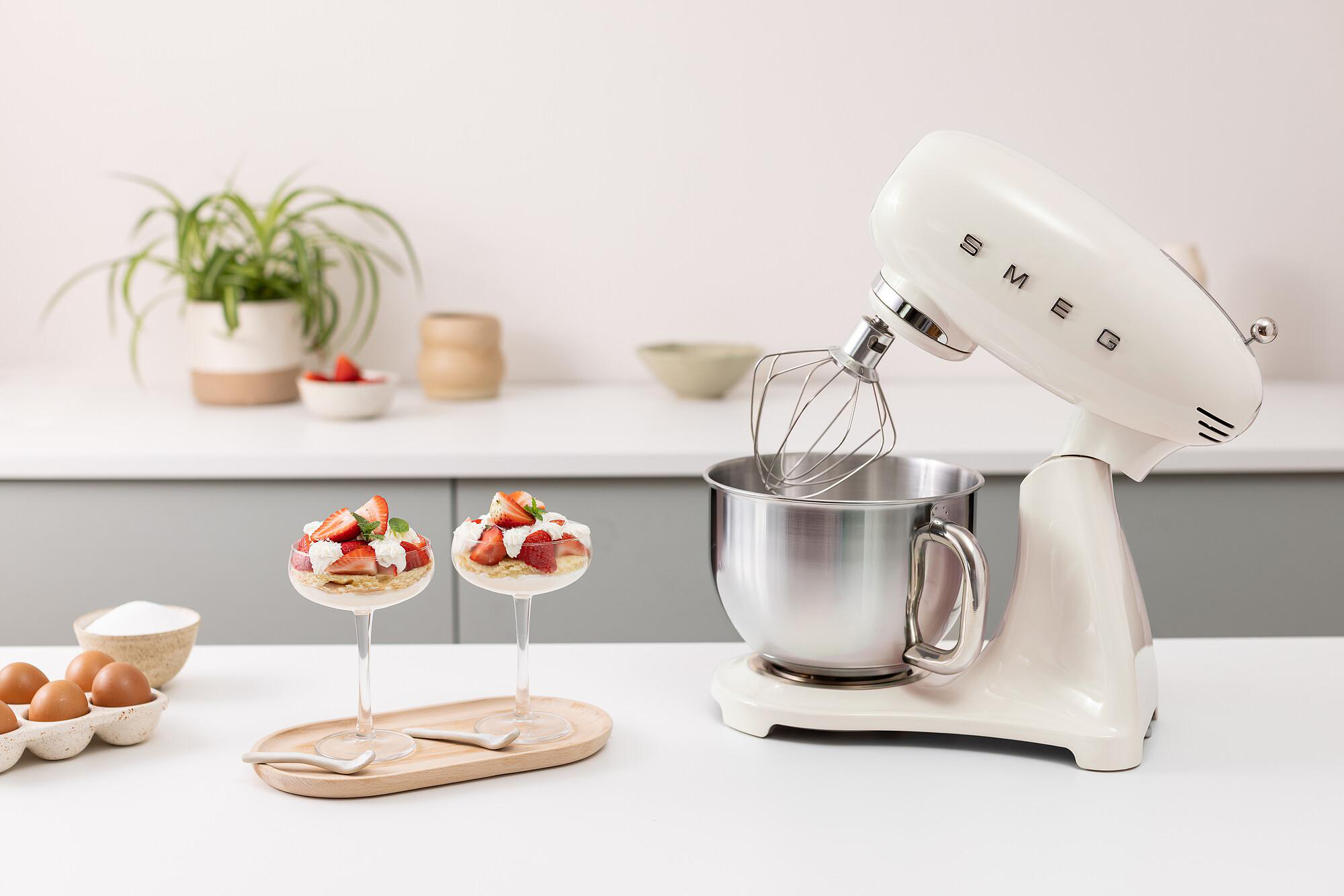Ein cremefarbener Mixer neben Desserts in Gläsern. Erdbeeren und Schlagsahne bedecken sie. Eier und Zucker sind vorhanden.