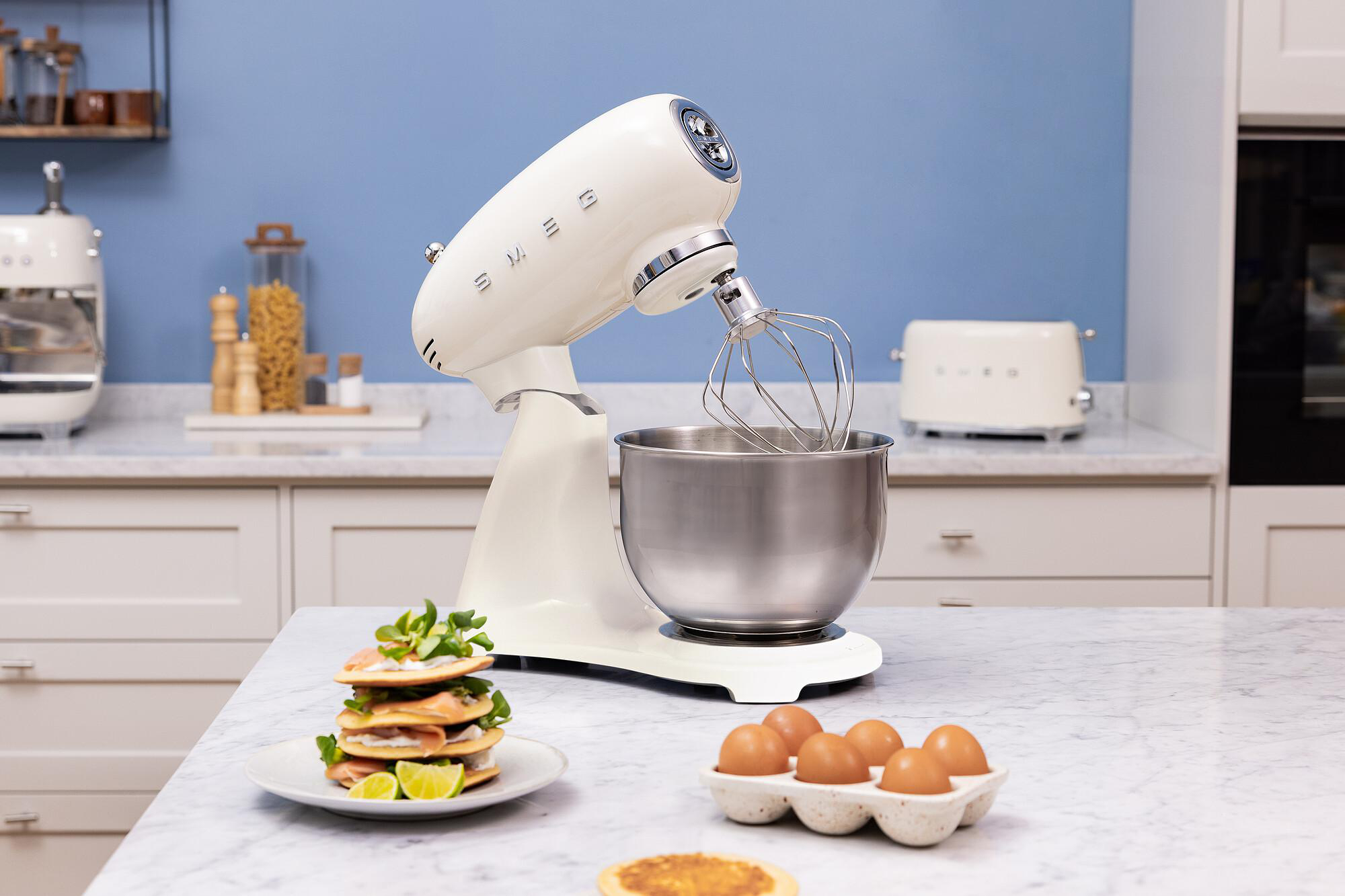 Ein cremefarbener Küchenmixer mit einer Edelstahlschüssel auf einer Marmorarbeitsplatte. Eier, Snacks und ein Toaster sind ebenfalls zu sehen.