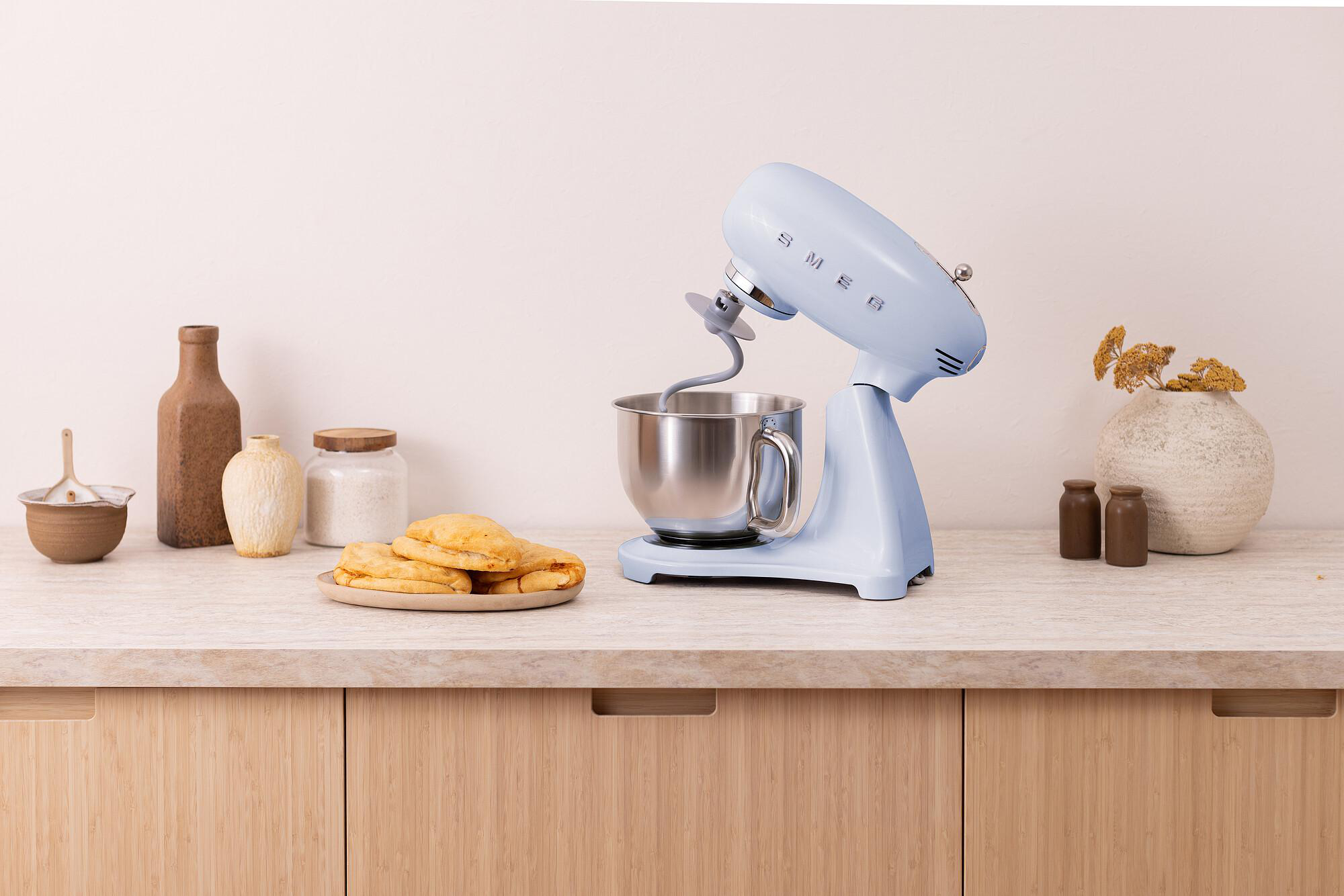 Ein hellblauer SMEG-Küchenmixer steht auf einer Theke mit anderen Küchenartikeln. Gebäck liegt auf einem Teller. Braune Flaschen und Gläser sind vorhanden.