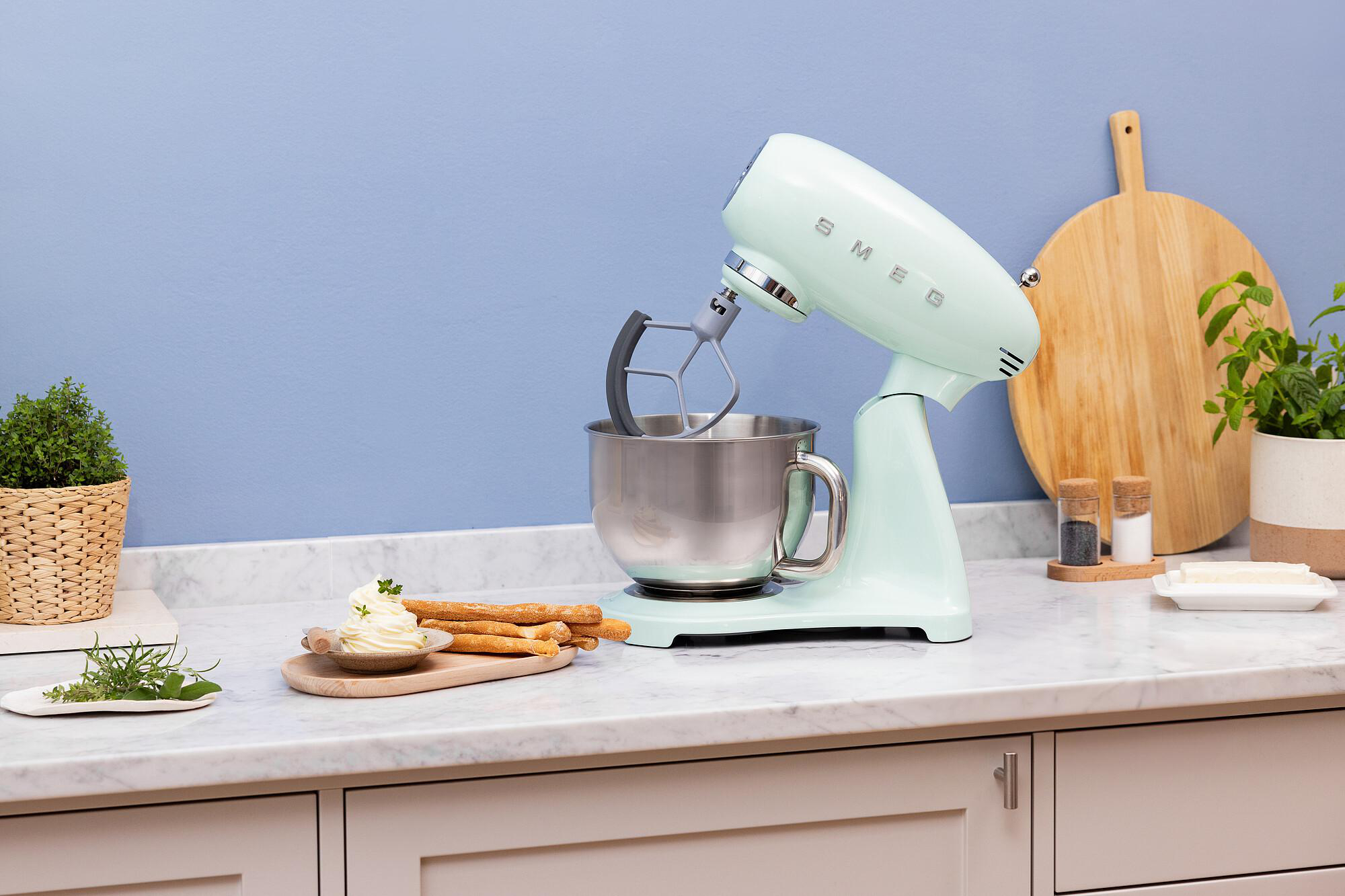 Ein mintgrüner Küchenmixer steht auf einer Marmorarbeitsplatte. Brotstangen, Butter und Pflanzen sind ebenfalls zu sehen. Im Hintergrund ist eine blaue Wand.