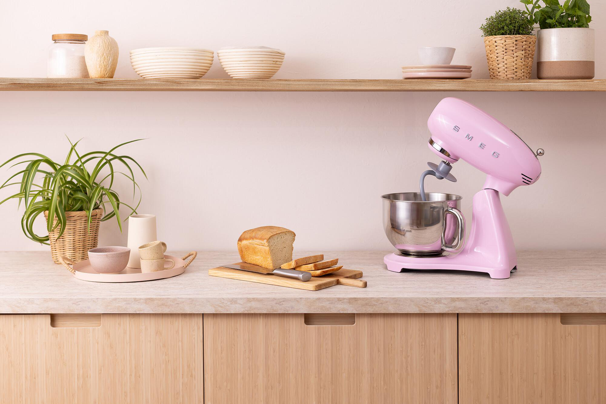 Ein rosa Mixer steht auf einer Arbeitsplatte neben geschnittenem Brot und einem Schneidebrett. Regale darüber halten Schüsseln und Pflanzen.