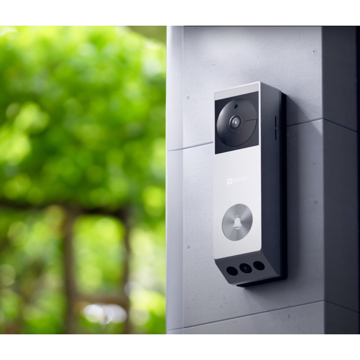 A silver doorbell camera is mounted on a gray wall. Green foliage is blurred in the background.