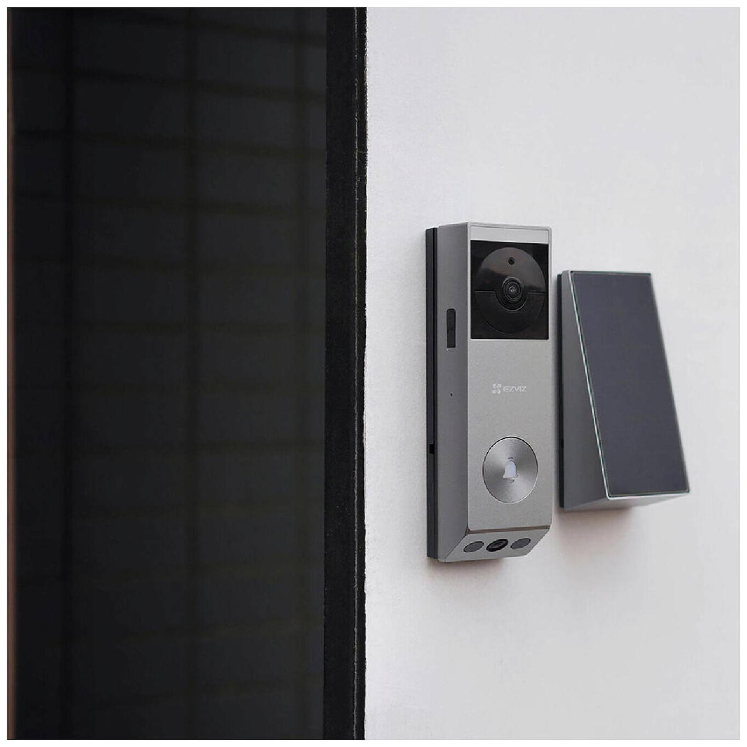 A silver doorbell with a camera is mounted on a white wall next to a dark door frame. A matching speaker is also present.