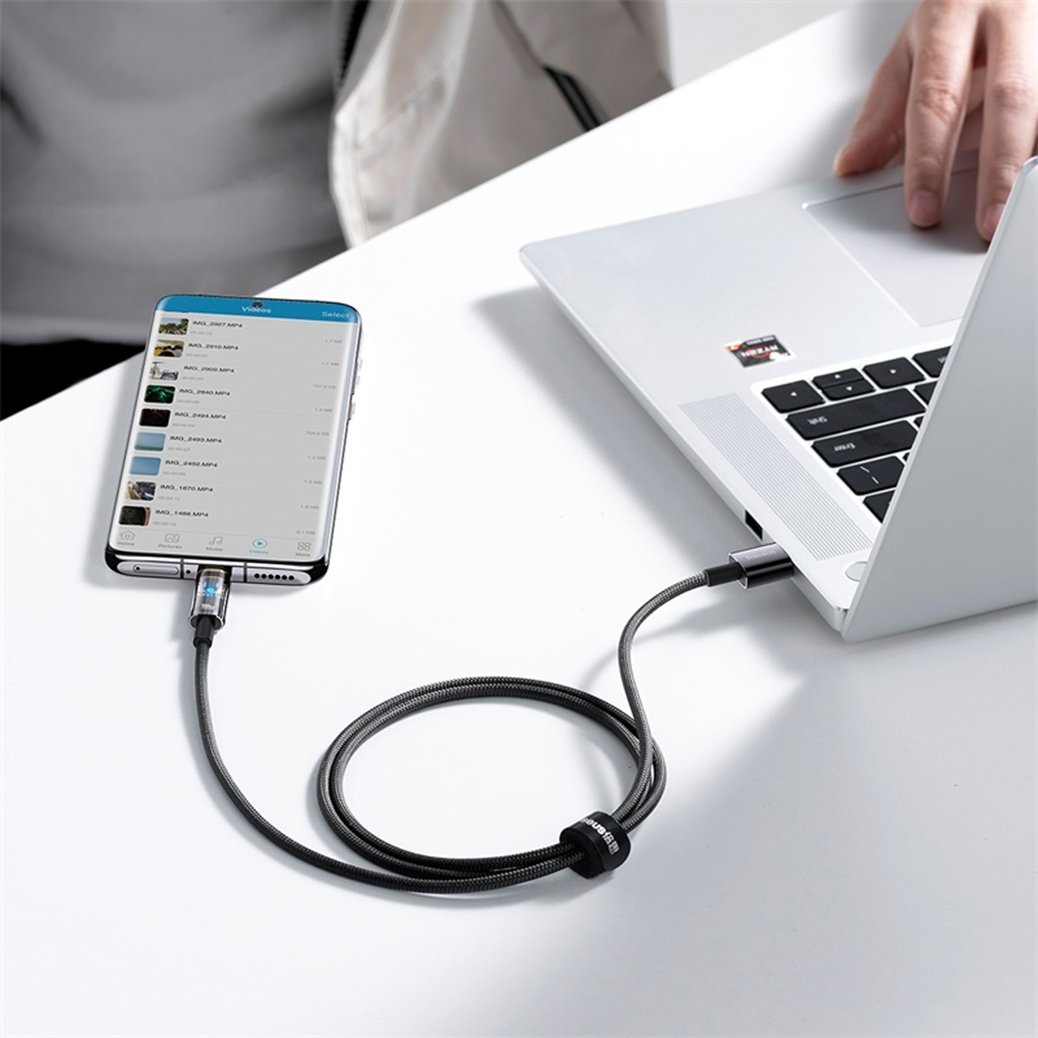 Un teléfono está conectado a una computadora portátil mediante un cable de carga. El teléfono muestra una lista de videos y la mano de una persona está cerca de la computadora portátil.