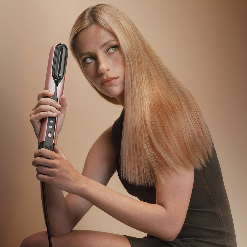 Una mujer sostiene una plancha de pelo rosa. Tiene cabello rubio y viste un vestido oscuro. Fondo beige.