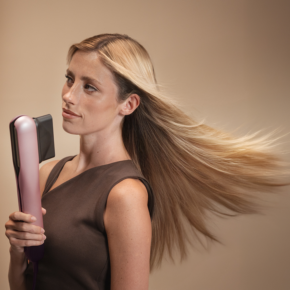 Mujer con cabello rubio ondeante usando una plancha de pelo rosa y negra, top marrón, fondo neutro.
