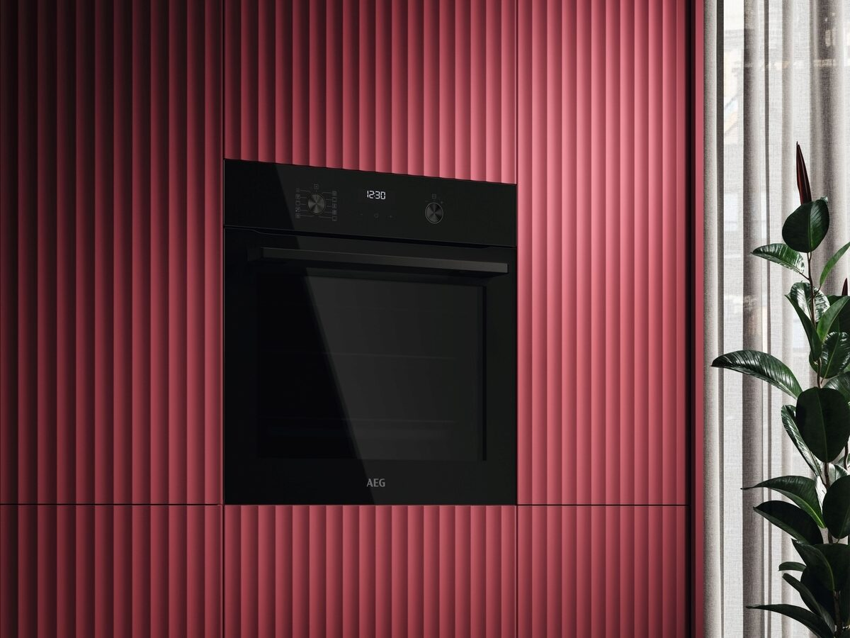 Un horno negro incorporado en un armario rojo texturizado. Se muestra la hora 12:30. Cortinas y una planta en el fondo.
