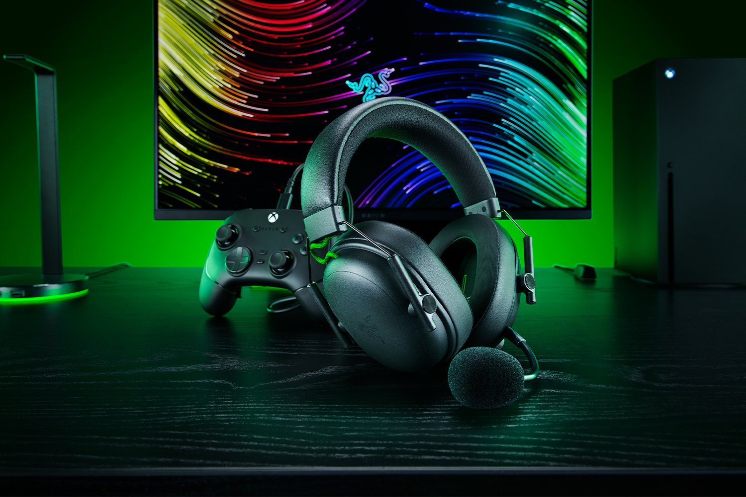 Gaming-Setup: Headset, Controller, Monitor mit buntem Hintergrund und Xbox. Dunkler Schreibtisch mit grüner Beleuchtung.