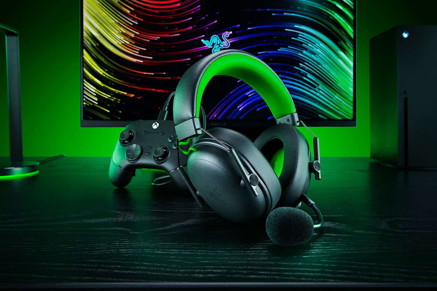 Schwarzes und grünes Gaming-Headset, Controller und Konsole auf einem schwarzen Schreibtisch vor einem bunten Monitor.
