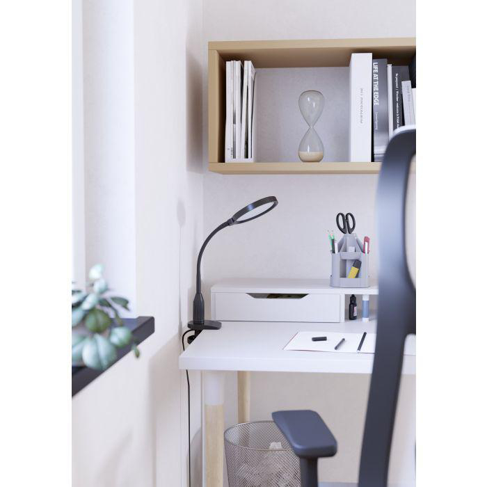 Ein Schreibtisch mit Lampe, Stifthalter und Regal mit Büchern und einer Sanduhr. Ein Stuhl ist im Vordergrund.