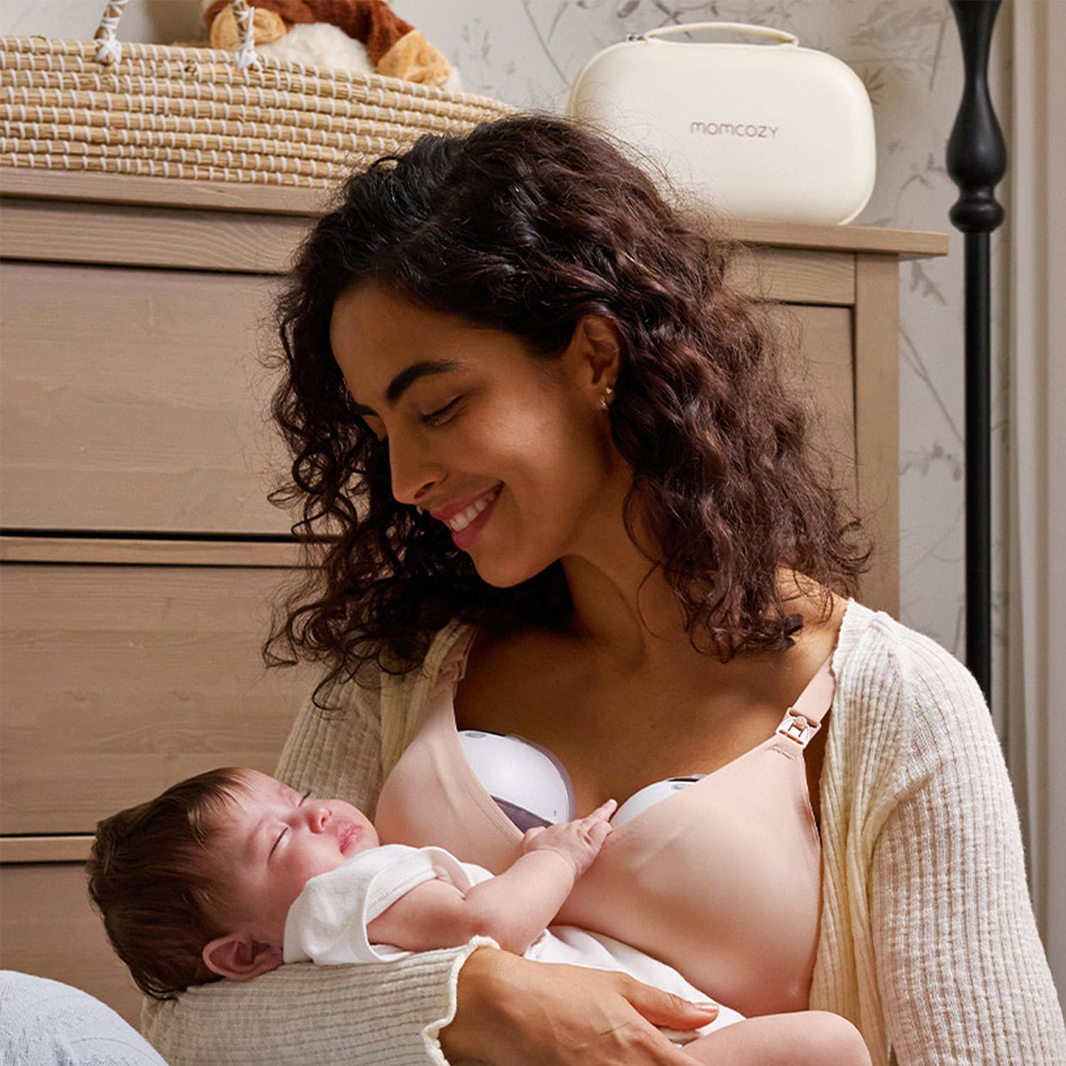 Eine lächelnde Frau hält ein Baby. Sie trägt einen Still-BH und eine weiße Strickjacke. Eine Milchpumpe ist angebracht.