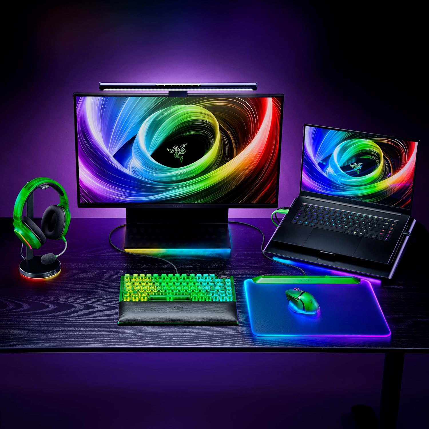 Gaming setup with a monitor, laptop, keyboard, mouse, headset, and mousepad, all with colorful LED lighting on a desk.
