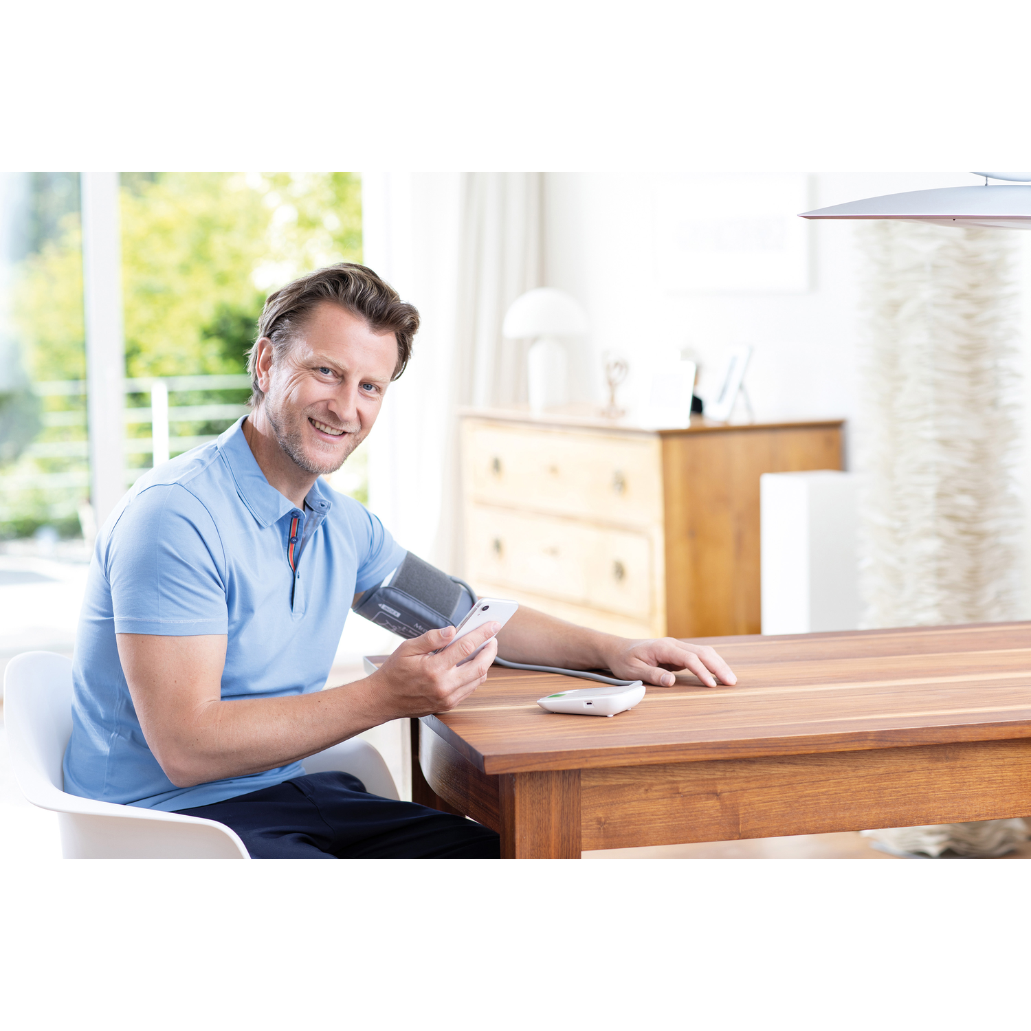 Man in blauw shirt controleert bloeddruk met apparaat op arm, zittend aan houten tafel. Witte stoel links.