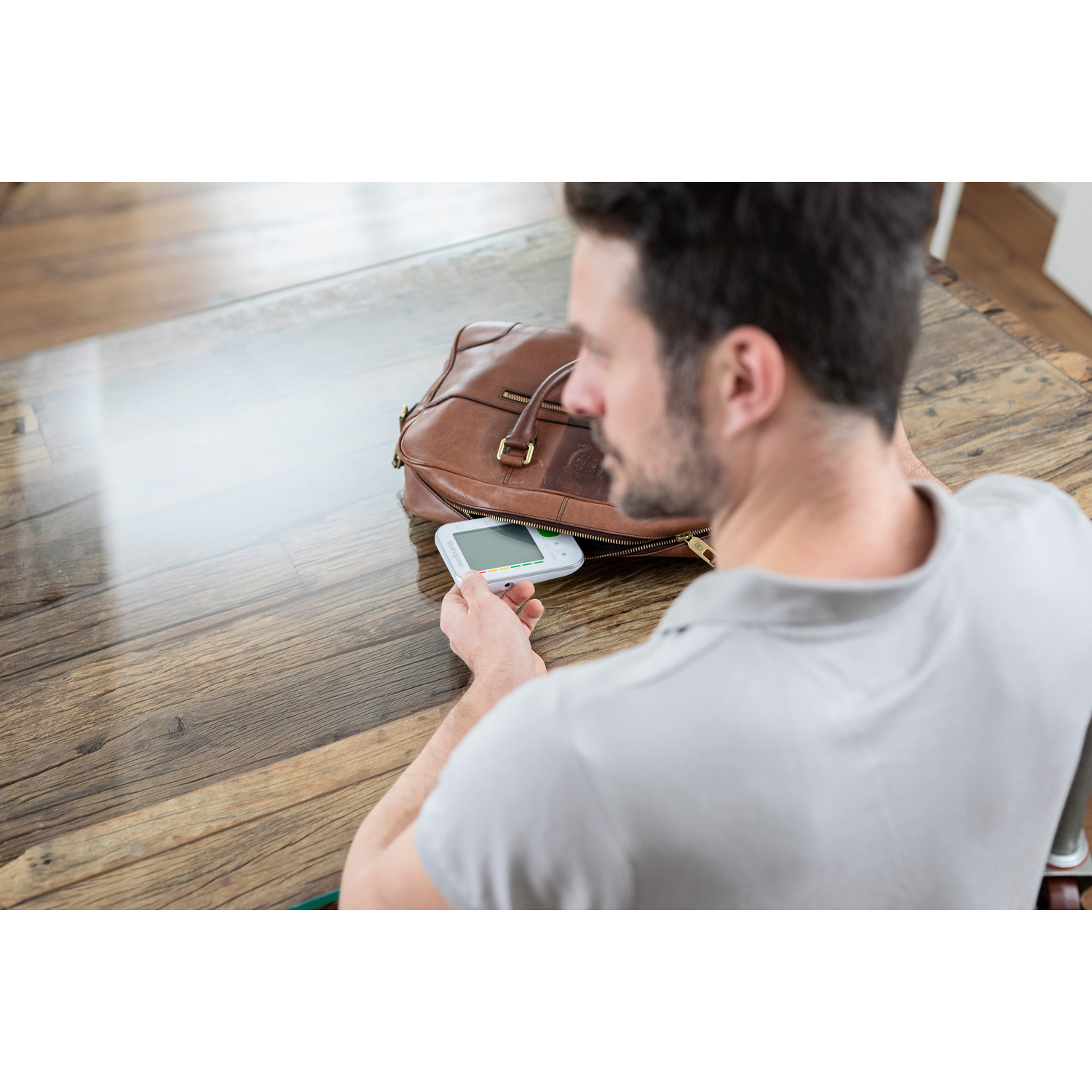 Een man in een grijs overhemd houdt een wit apparaat vast naast een bruine leren tas op een houten tafel.