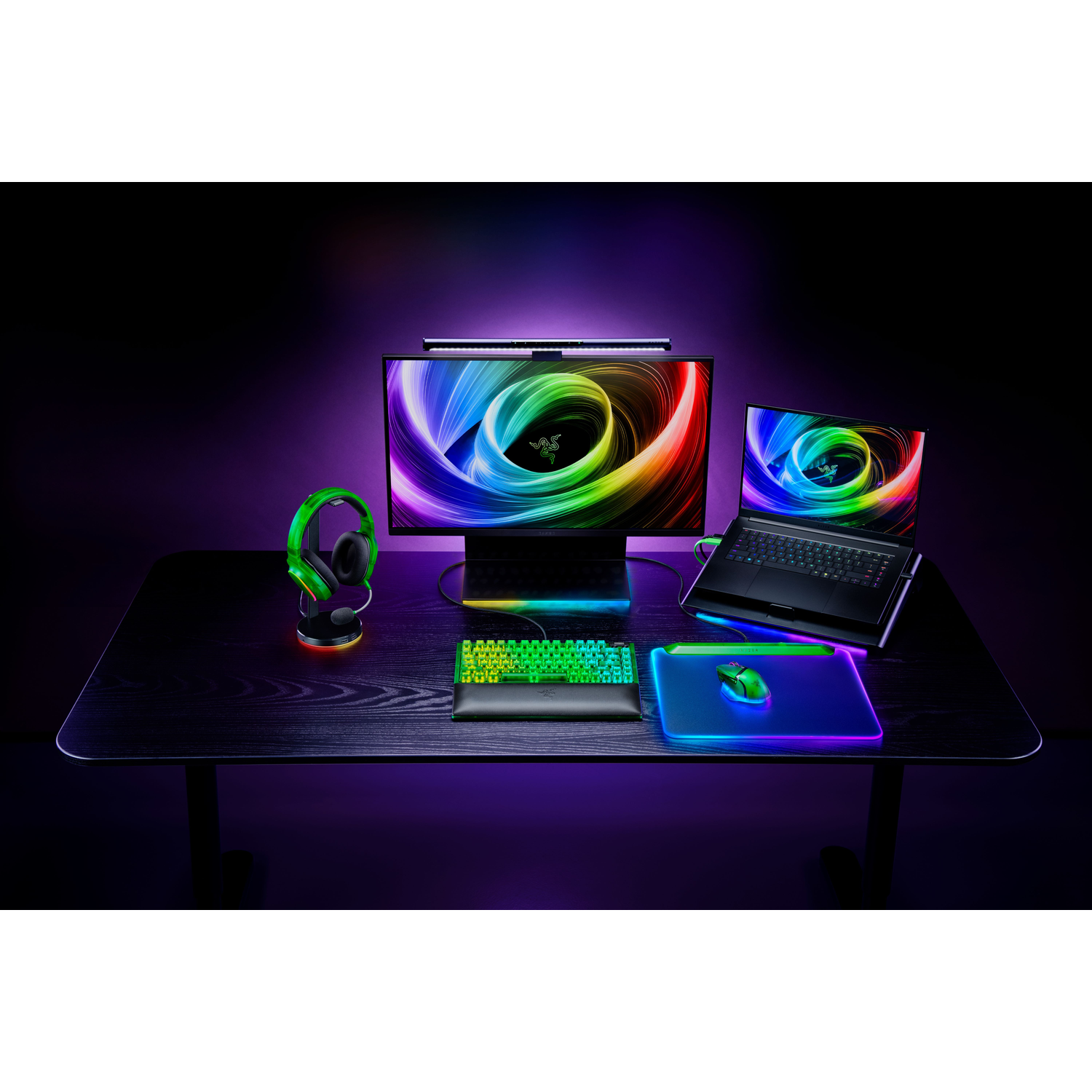 Gaming setup met monitor, laptop, toetsenbord, muis en headset, allemaal verlicht met regenboogkleuren op een zwart bureau.