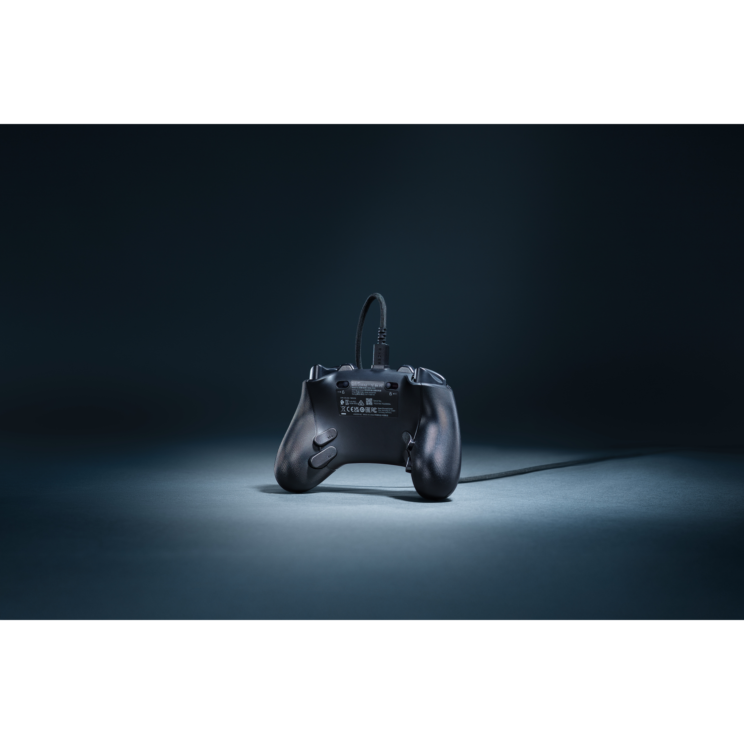 A black gamepad with a cable lies on a surface. The gamepad is angled towards the viewer, the background is dark.