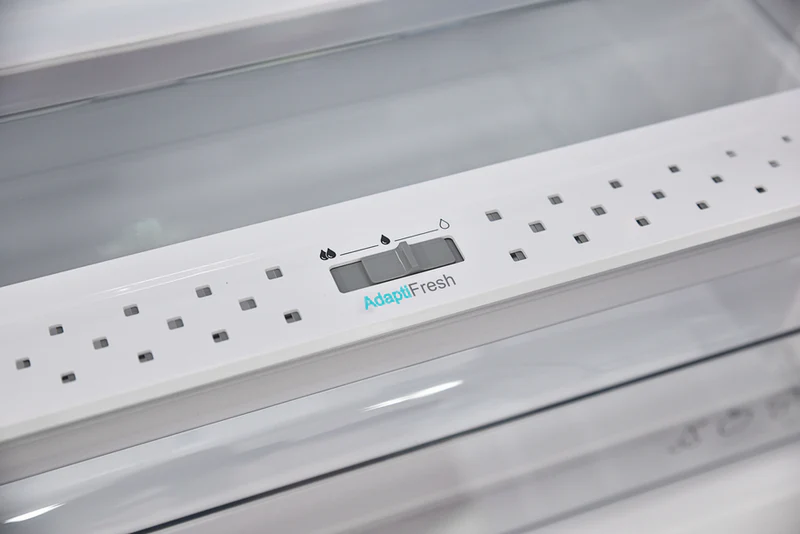 Egy hűtőszekrény belsejében egy fehér vezérlőpanel 'AdaptiFresh' felirattal és egy csúszkával. A háttér egy átlátszó polc.