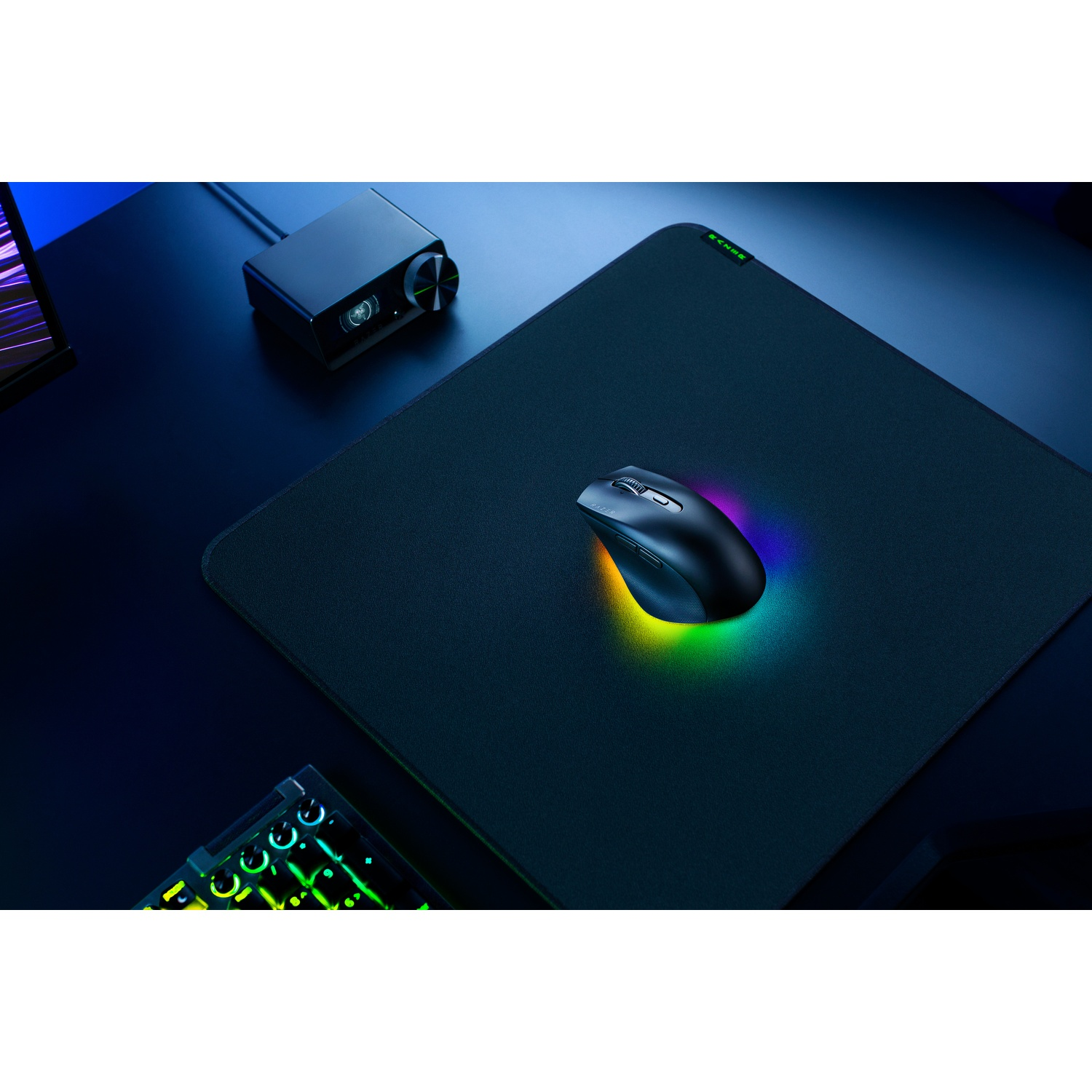 Une souris noire sur un tapis de souris noir avec éclairage RVB. À gauche, un boîtier de commande noir et en bas à gauche, un clavier.