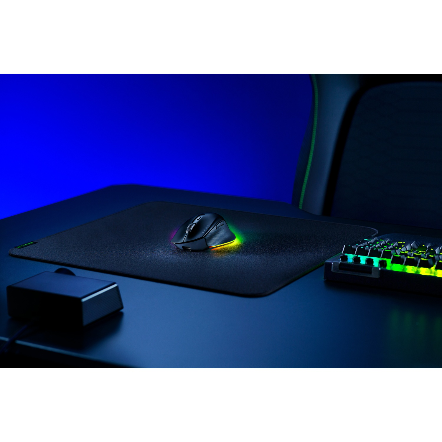 Une souris noire sur un tapis, un clavier et un boîtier noir sur un bureau bleu foncé. Une chaise est visible en arrière-plan.