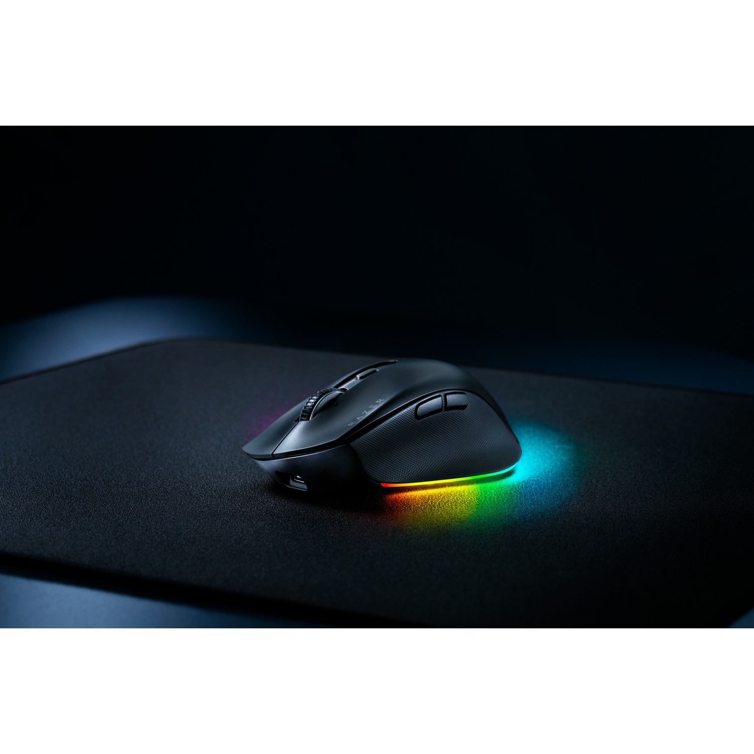 Une souris Razer noire repose sur un tapis de souris noir. La souris a des lumières LED colorées sur le bord inférieur. Fond bleu foncé.