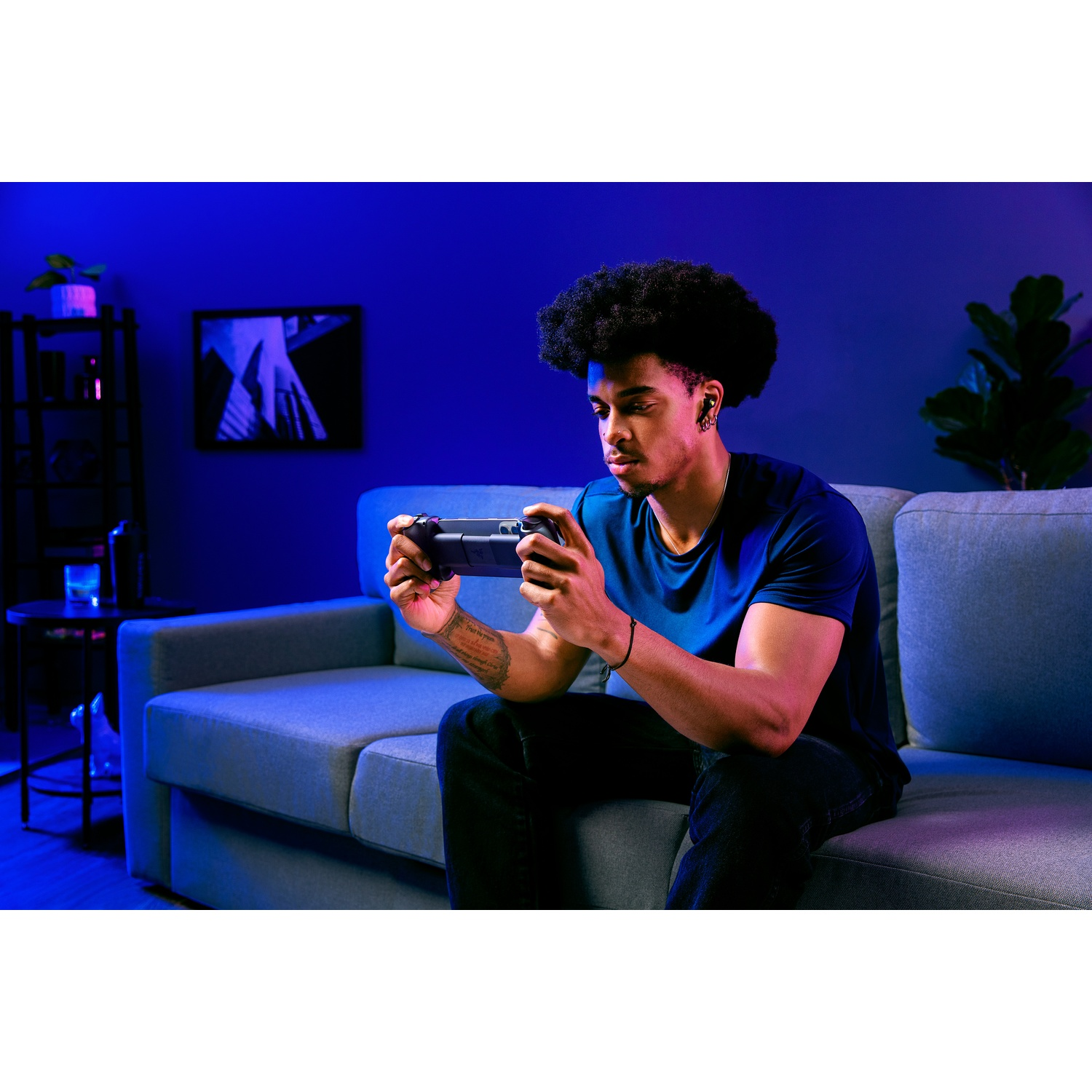 Een man zit op een bank en speelt met een handheld console. Hij draagt een blauw shirt en heeft een koptelefoon. De kamer is zwak verlicht.